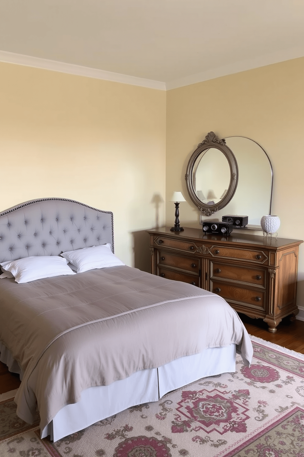 A cozy bedroom in a one-bedroom apartment featuring a plush queen-sized bed with a tufted headboard in a soft gray fabric. A vintage wooden dresser with ornate detailing sits against the wall, complemented by a large round mirror with a distressed finish above it. The walls are painted in a warm cream color, creating a welcoming atmosphere. A stylish area rug with intricate patterns lies beneath the bed, adding texture and warmth to the space.