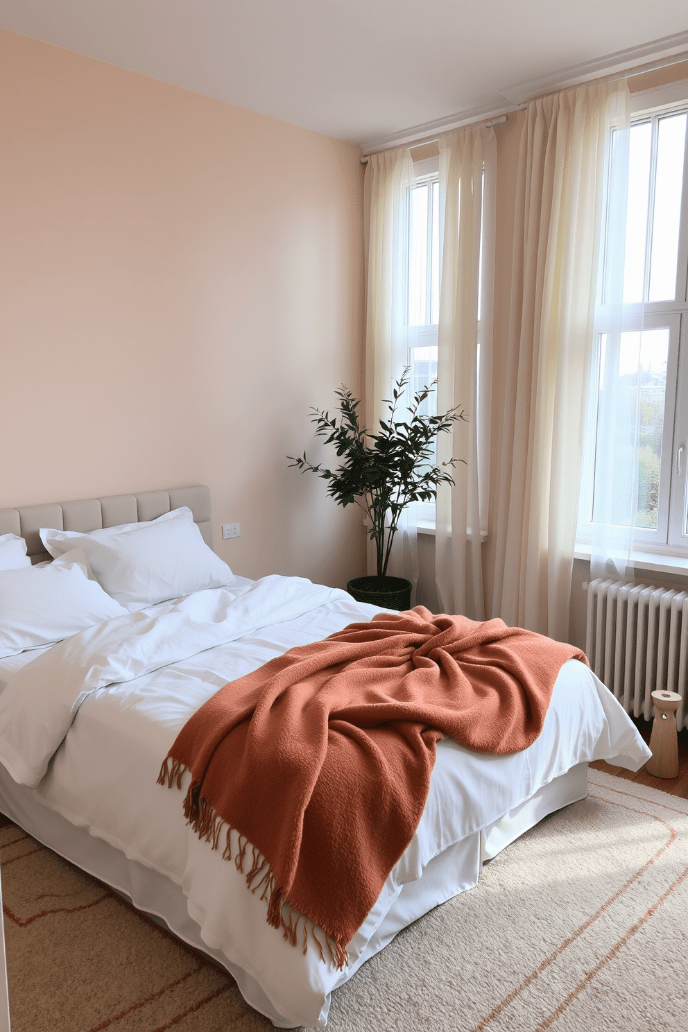 A cozy apartment bedroom featuring a plush bed dressed in soft linens and a warm throw blanket draped casually across the foot. The walls are painted in a soothing pastel color, and large windows allow natural light to fill the space, complemented by sheer curtains.