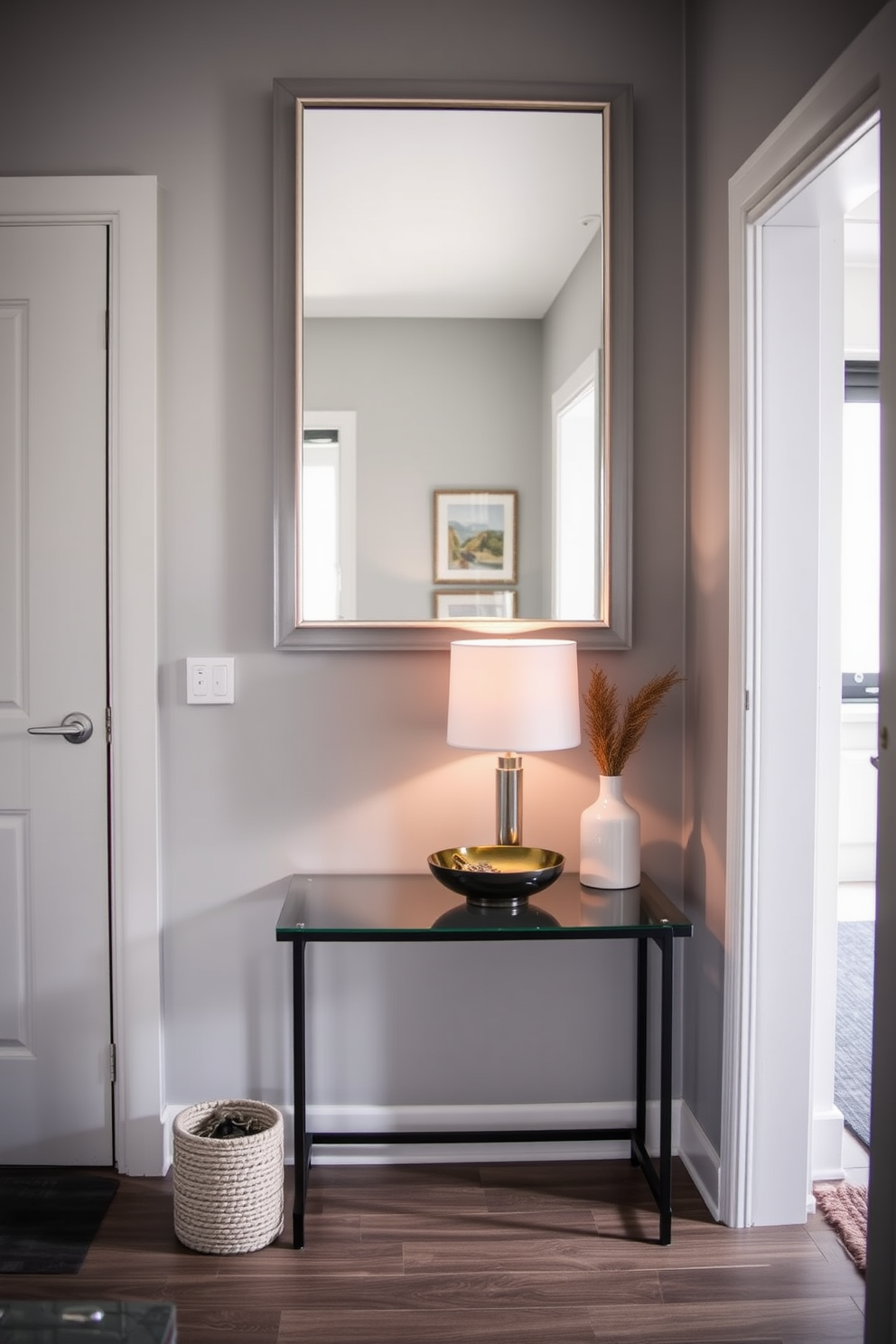 A stylish apartment entryway features a small console table against the wall, adorned with a decorative lamp and a stylish bowl for keys. The walls are painted in a soft gray tone, and a large mirror hangs above the table, reflecting natural light into the space.