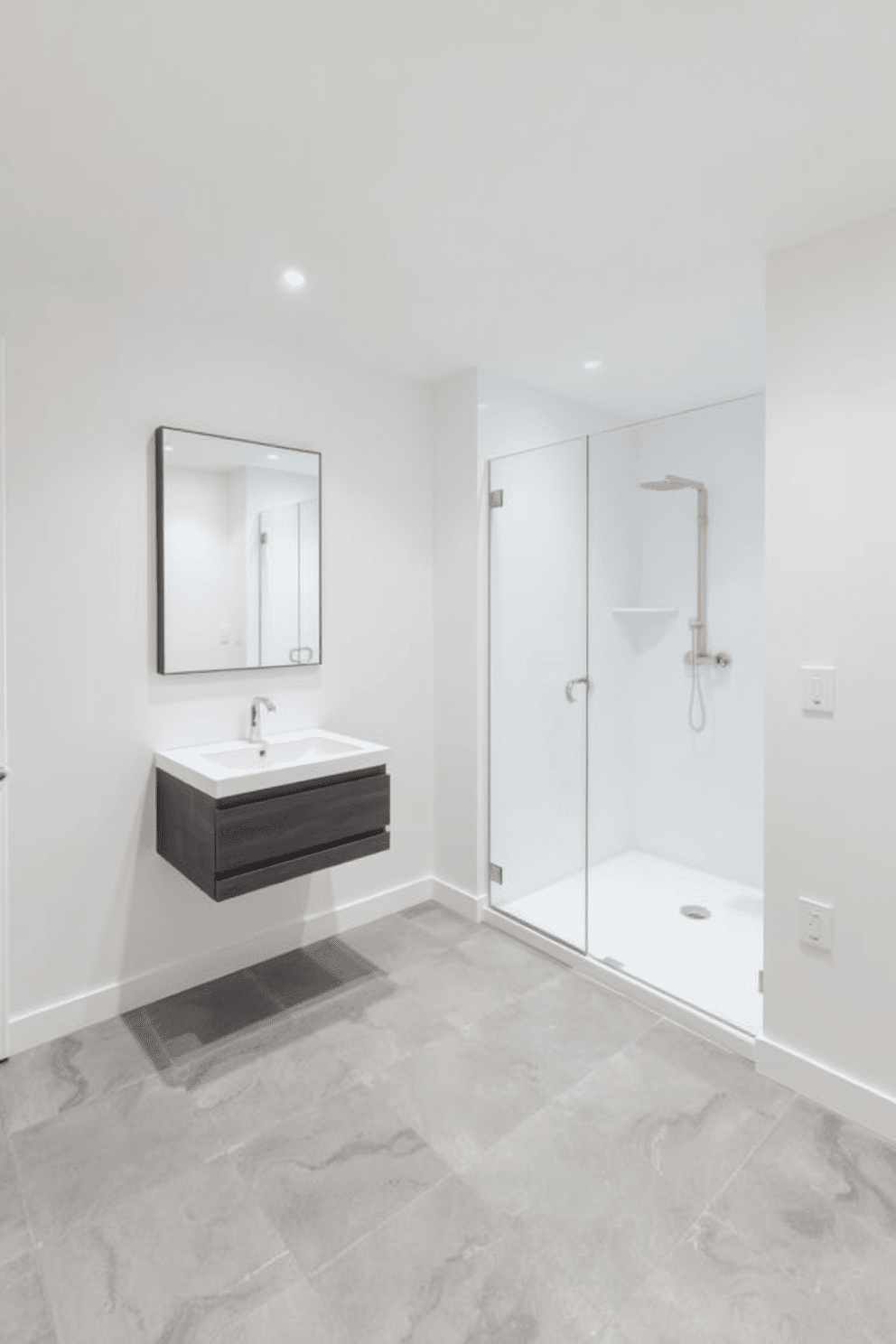 A minimalist basement bathroom featuring sleek lines and a spacious layout. The walls are painted in a soft white, and the floor is adorned with large gray tiles for a modern touch. A floating vanity with a single sink is positioned against one wall, complemented by a large frameless mirror above it. The shower area is enclosed with clear glass, showcasing a rainfall showerhead and simple, elegant fixtures.