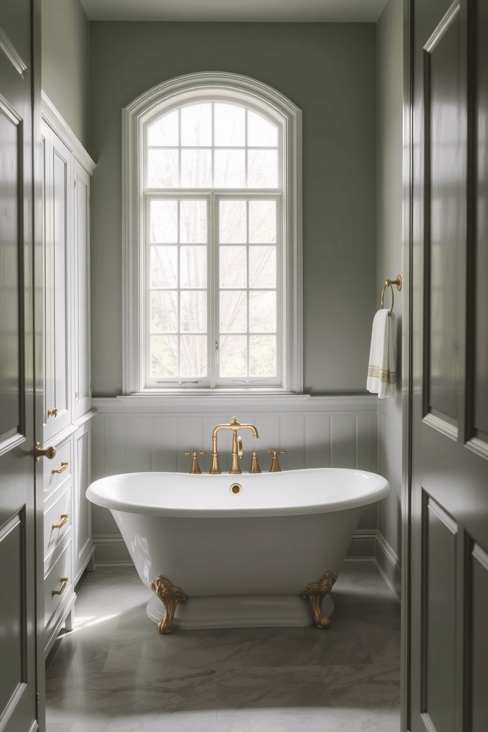 A serene bathroom space featuring muted sage green walls that evoke a calming natural atmosphere. The design incorporates white cabinetry and brass fixtures to enhance the tranquility of the setting. The floor is adorned with light gray tiles that complement the sage green tones. A freestanding bathtub sits elegantly beneath a large window, allowing natural light to flood the room.