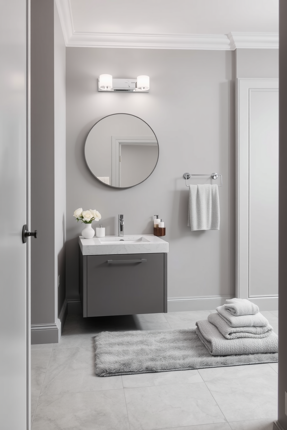 A chic powder room featuring a monochromatic color scheme that exudes sophistication. The walls are painted in a soft gray, complemented by a sleek gray vanity with a polished stone countertop. Above the vanity, an elegant round mirror reflects the understated beauty of the space. Subtle lighting fixtures with a modern design illuminate the room, enhancing the serene atmosphere. The floor is adorned with large gray tiles that create a seamless look. A plush gray rug adds warmth underfoot, while minimalistic decor accents the refined aesthetic. Decorative elements include a simple vase with white flowers and neatly folded towels in varying shades of gray. The overall design promotes a sense of tranquility and elegance, making it the perfect retreat.