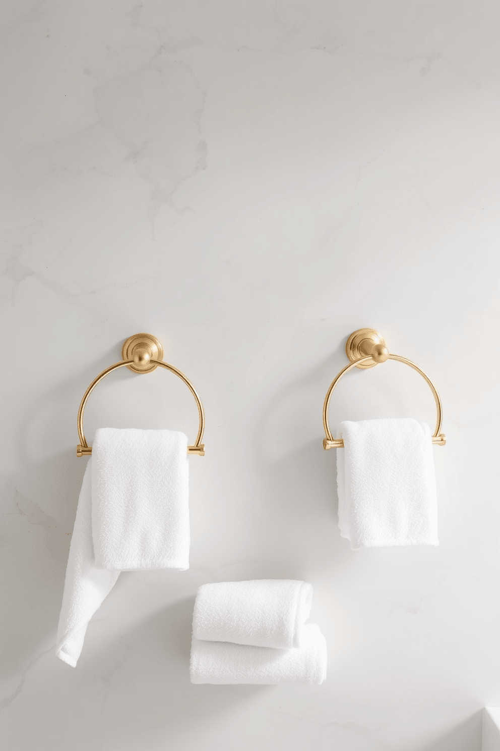 Elegant towel rings mounted on the wall in a luxurious bathroom setting. The towel rings are made of polished brass, complementing the soft color palette of the space. Each towel ring holds a plush white towel, neatly arranged for easy access. The backdrop features a serene light gray wall with subtle textures, enhancing the overall sophistication of the design.