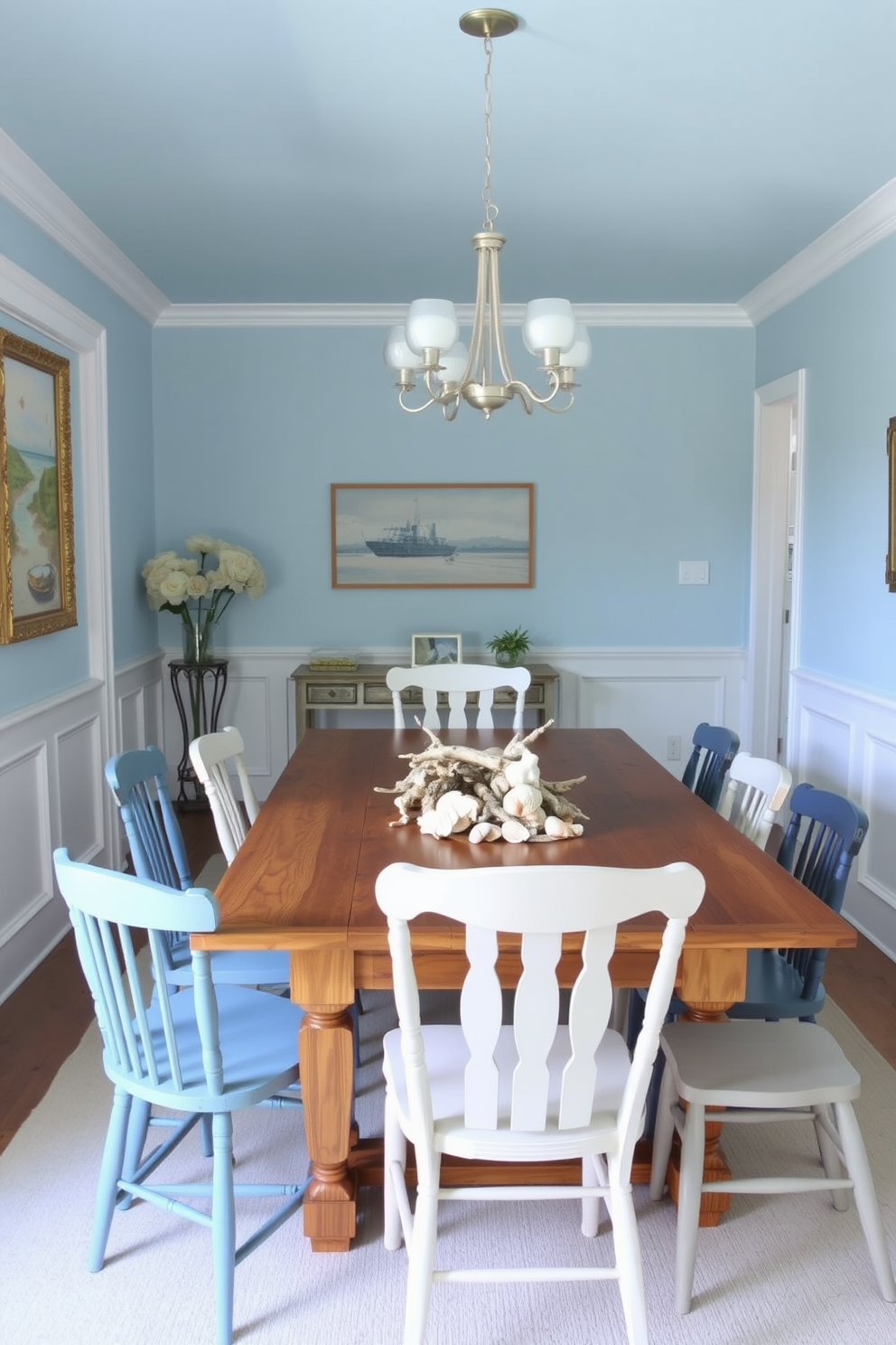 A coastal dining room featuring soft blue walls and white wainscoting. A large wooden dining table is surrounded by mismatched chairs in shades of blue and white, with a centerpiece of seashells and driftwood.