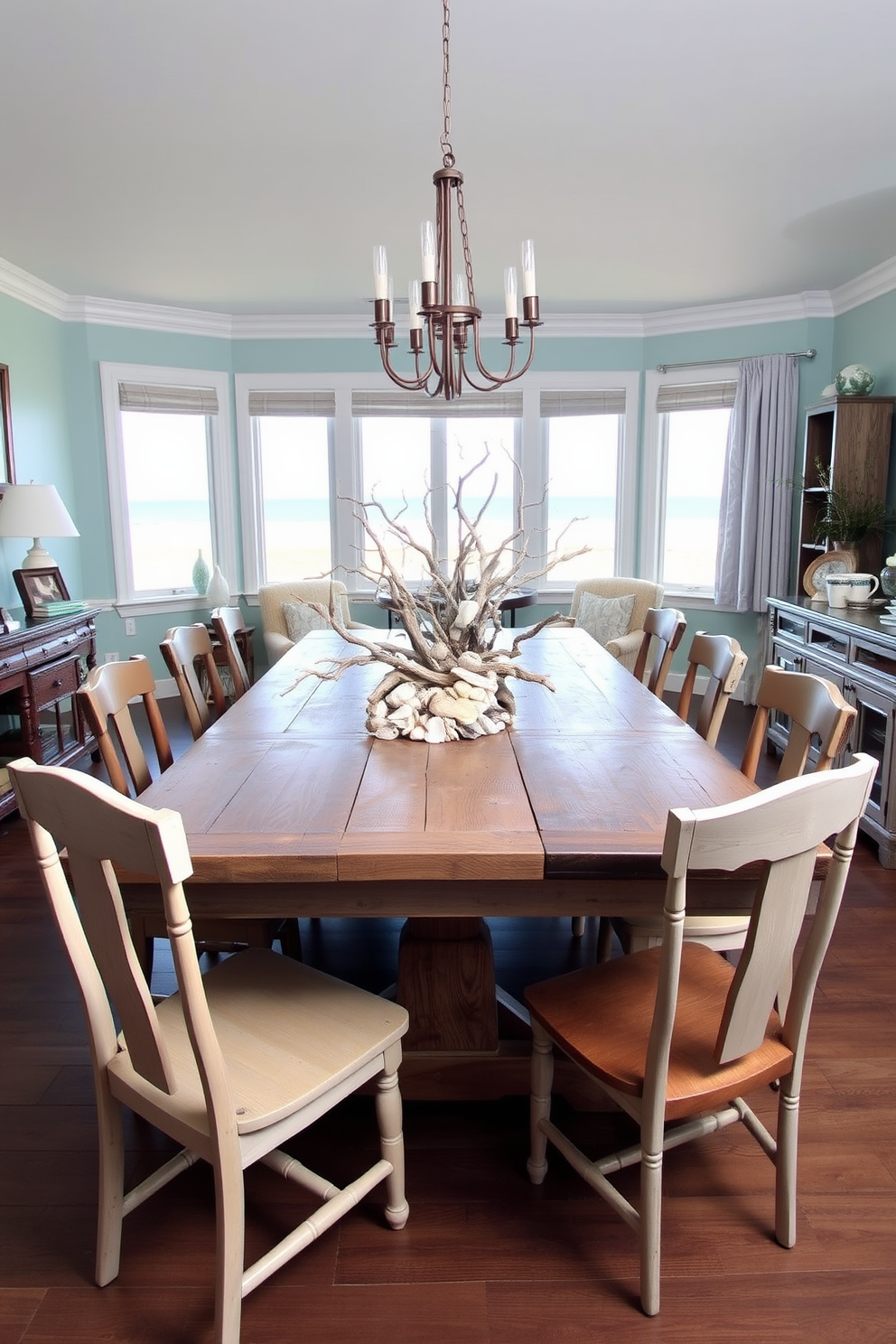 A beachy dining room setting features a large wooden table with a weathered finish surrounded by mismatched chairs. Centered on the table is a stunning centerpiece made of driftwood and an assortment of shells, evoking a coastal vibe. The walls are painted in a soft aqua hue, complementing the sandy beige tones of the decor. Large windows allow natural light to flood the space, showcasing ocean views and enhancing the relaxed atmosphere.