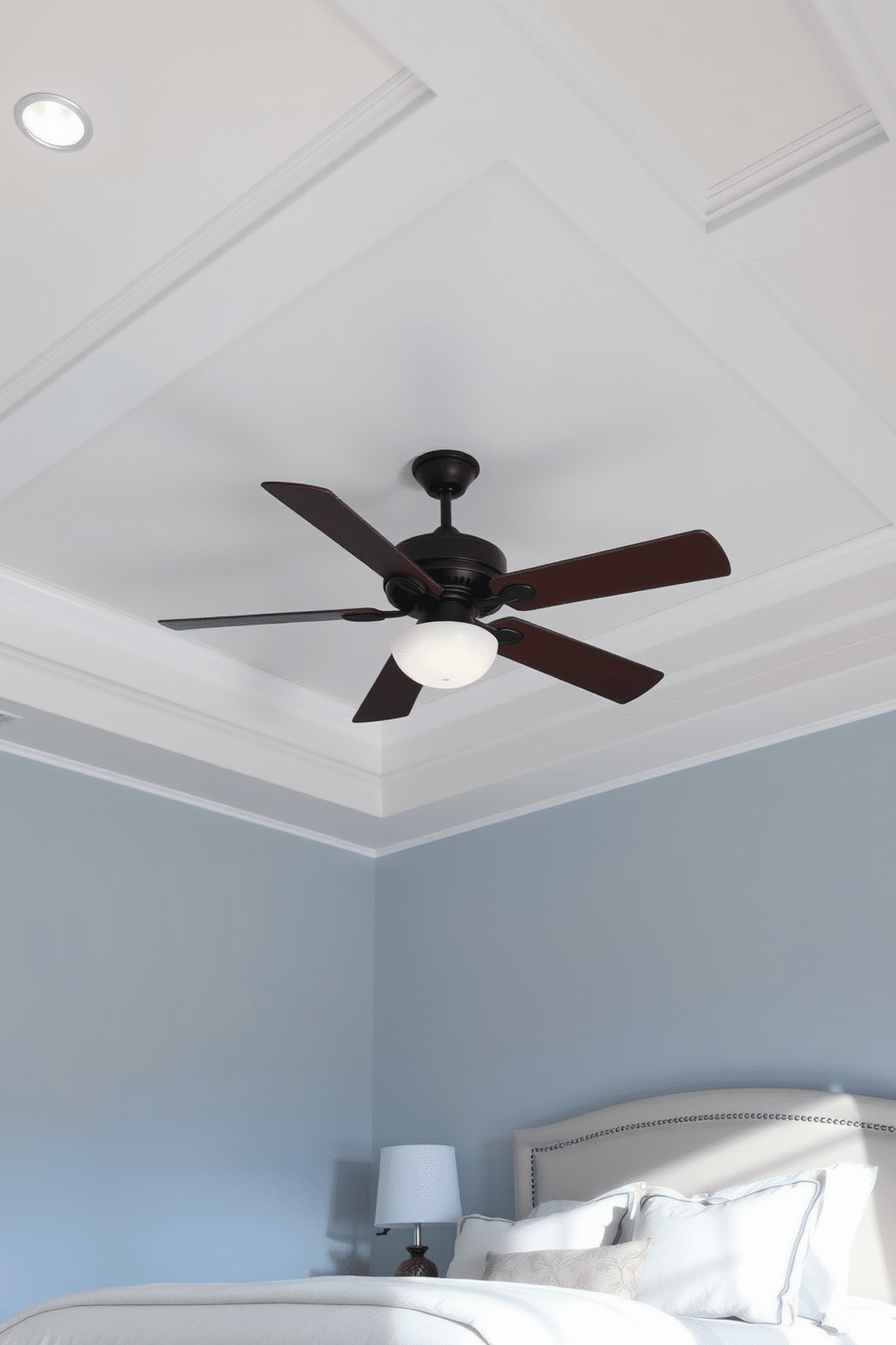 A serene bedroom featuring an integrated ceiling fan that complements the overall design. The ceiling is adorned with soft recessed lighting and a subtle coffered pattern, enhancing the room's elegance. The walls are painted in a calming light blue hue, providing a tranquil backdrop for relaxation. A plush king-sized bed with layered bedding sits beneath the fan, creating a cozy and inviting atmosphere.