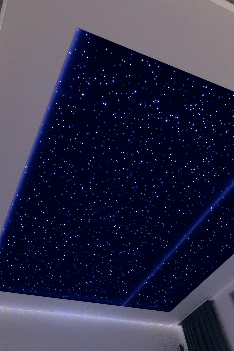 A starlight ceiling creates a dreamy atmosphere in the bedroom. Tiny LED lights are embedded in a dark blue fabric that mimics a night sky, twinkling softly like stars. The ceiling design features a subtle glow that enhances the overall ambiance of the room. Soft, recessed lighting around the edges of the ceiling adds warmth and depth to the space.