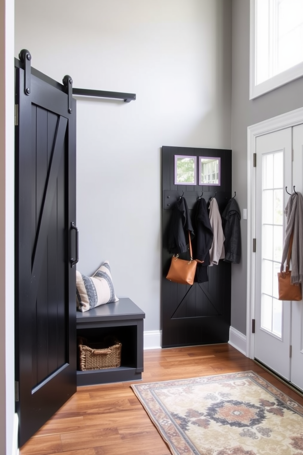 A stylish mudroom featuring a black sliding barn door that adds a rustic charm. The walls are painted in a soft gray hue, complemented by a bench with built-in storage beneath. On the right side, a row of hooks is mounted for coats and bags, while a large area rug in a geometric pattern anchors the space. Natural light floods in through a nearby window, enhancing the inviting atmosphere.