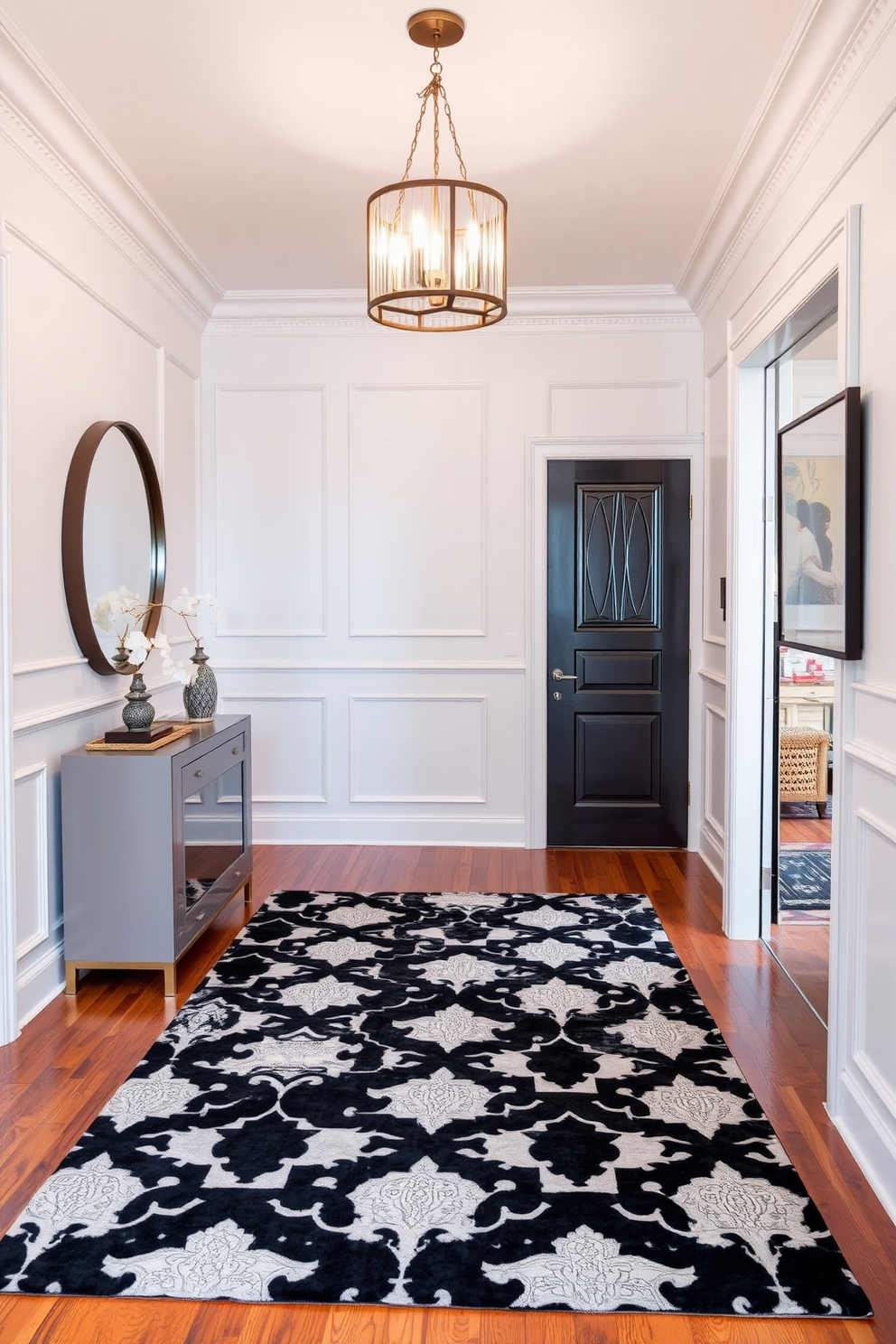 A stylish foyer featuring a black and white patterned area rug that adds visual interest to the space. The walls are adorned with elegant wainscoting, and a statement chandelier hangs from the ceiling, casting a warm glow. To the left, a sleek console table with a glossy finish holds decorative items and a large round mirror. The flooring is a rich hardwood, providing a stunning contrast to the rug and enhancing the overall sophistication of the foyer.