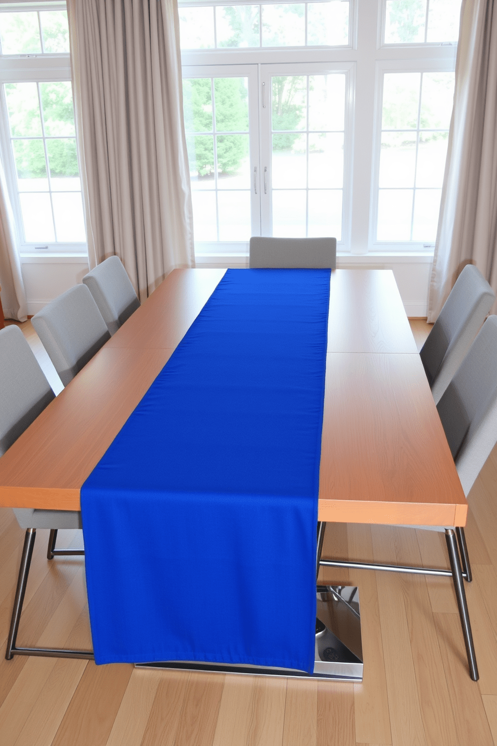 A sapphire blue table runner elegantly drapes across a modern dining table, contrasting beautifully with the warm wooden surface beneath. Surrounding the table are sleek, contemporary chairs upholstered in a light gray fabric, creating a harmonious balance of color and texture. The dining area features large windows that allow natural light to flood in, highlighting the vibrant blue of the table runner. Soft, neutral curtains frame the windows, adding a touch of sophistication to the overall design.