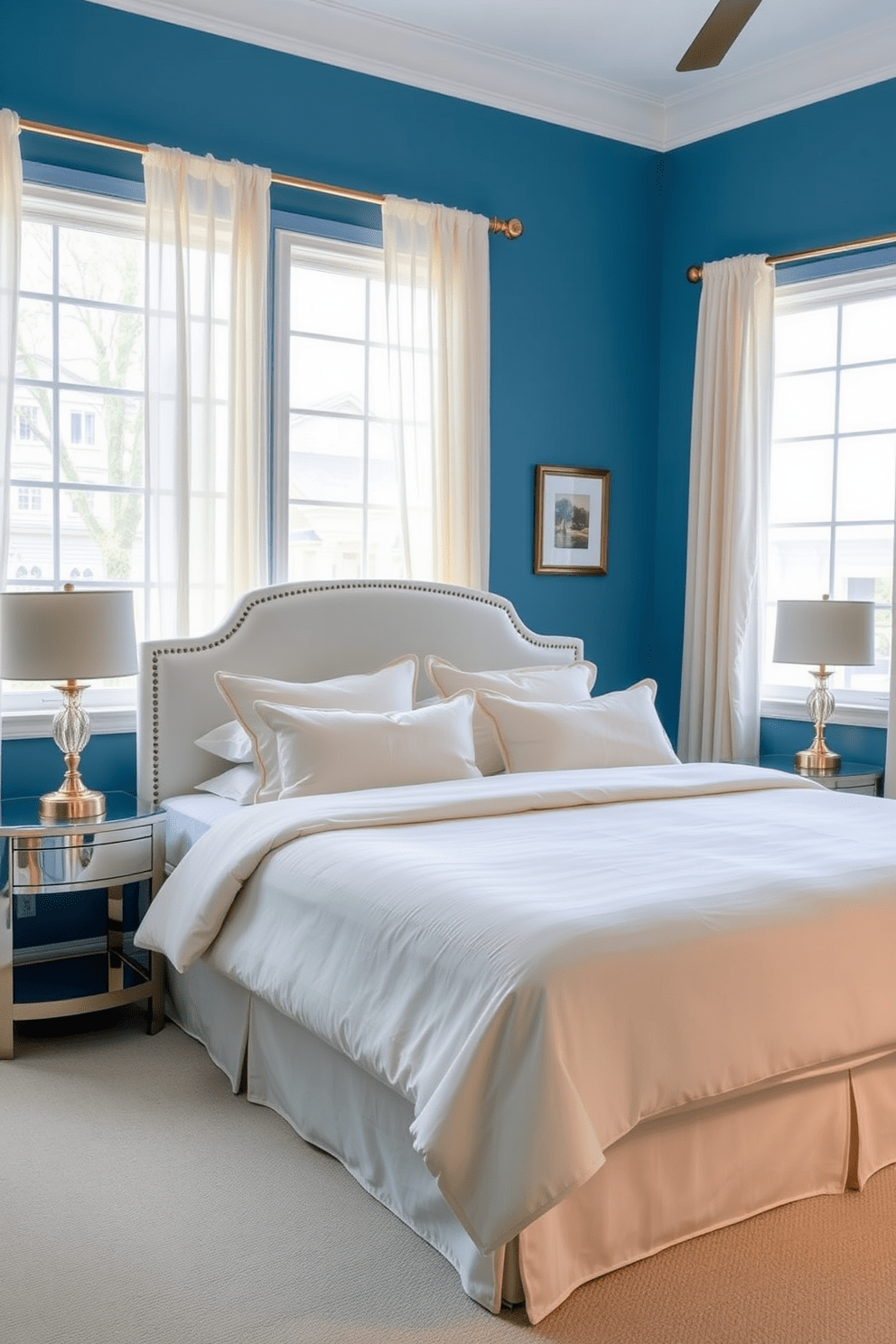 A serene blue bedroom setting. The walls are painted in a soft blue hue, complemented by cream-colored bedding on a plush king-size bed. A pair of elegant nightstands flanks the bed, each topped with stylish lamps that cast a warm glow. Large windows draped with sheer cream curtains allow natural light to filter in, enhancing the tranquil atmosphere.