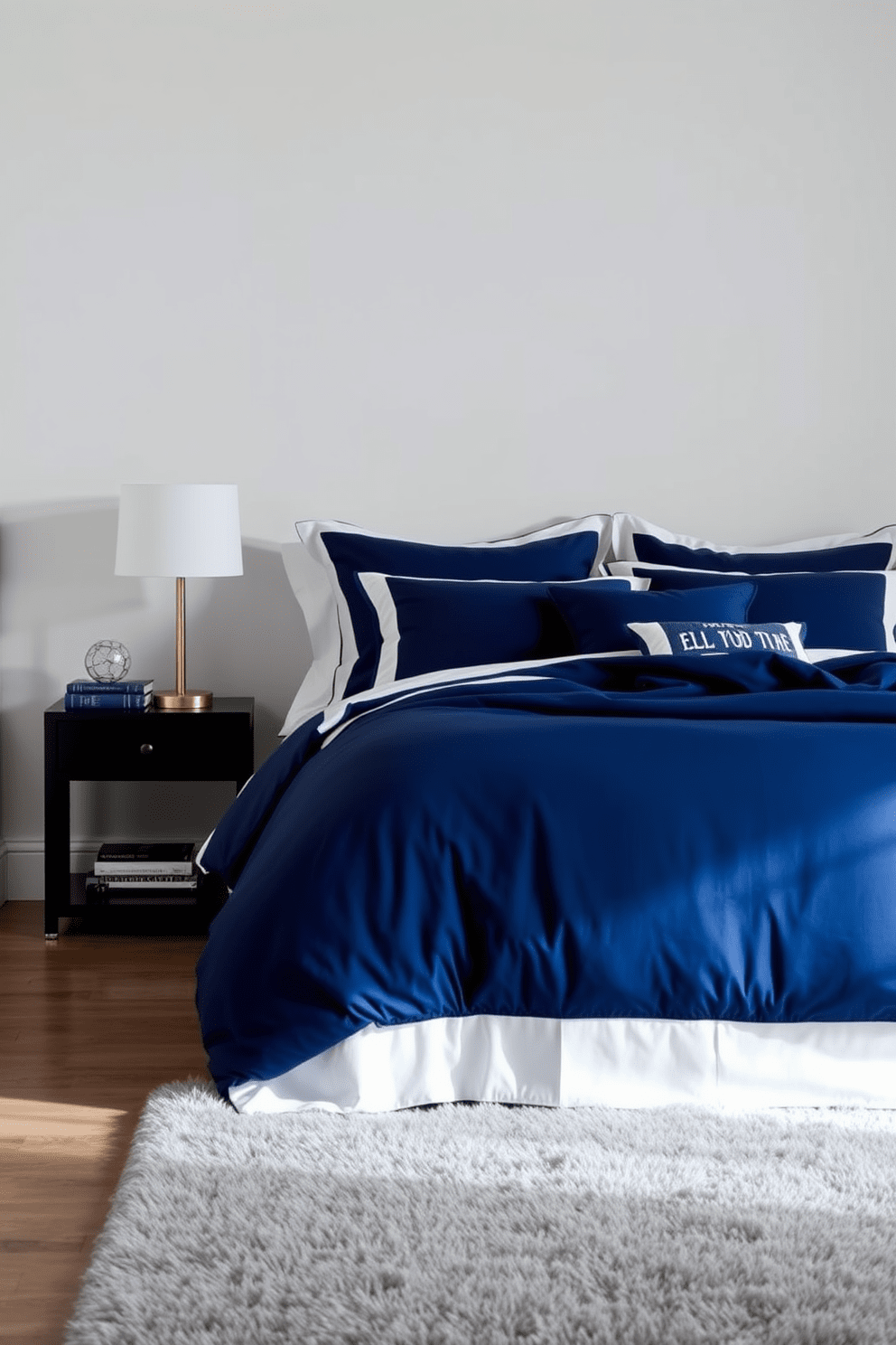 A serene bedroom featuring indigo blue bedding adorned with crisp white trim. The walls are painted in a soft neutral shade, creating a calming backdrop for the bold bedding. A stylish nightstand sits beside the bed, topped with a modern lamp and a few decorative books. The floor is covered with a plush area rug that complements the color scheme, adding warmth to the space.