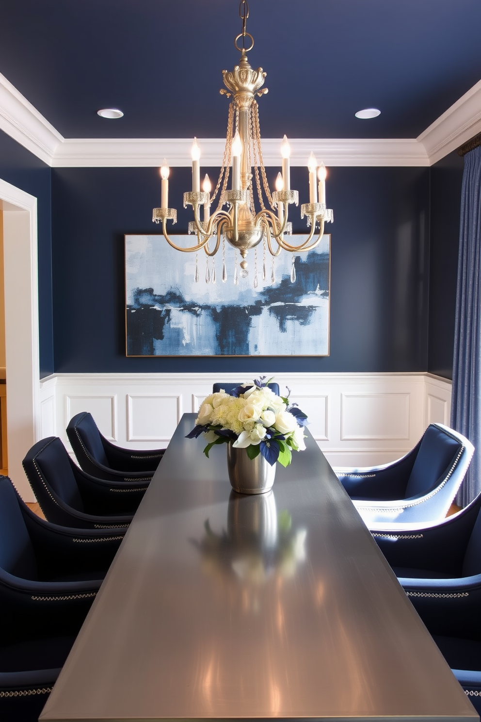 Elegant navy and silver theme Blue Dining Room Design Ideas. The dining room features a long rectangular table with a polished silver finish and navy upholstered chairs. Above the table, a stunning chandelier with silver accents hangs from the ceiling, casting a warm glow. The walls are painted a deep navy blue, complemented by white wainscoting that adds a touch of sophistication. A large abstract painting in shades of blue and silver adorns one wall, serving as a focal point. On the table, a simple yet elegant floral arrangement in silver and white completes the refined look.