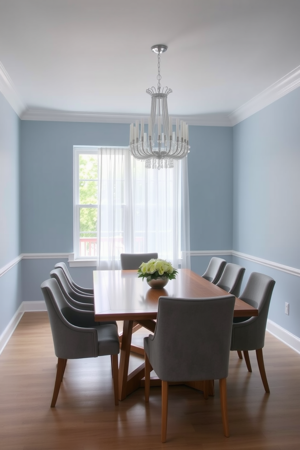 A serene dining room featuring a soft blue and gray color palette. The walls are painted in a light blue hue, complemented by gray upholstered dining chairs around a sleek wooden table. A large window allows natural light to flood the space, with sheer white curtains gently framing the view. A statement chandelier hangs above the table, adding an elegant touch to the overall design.