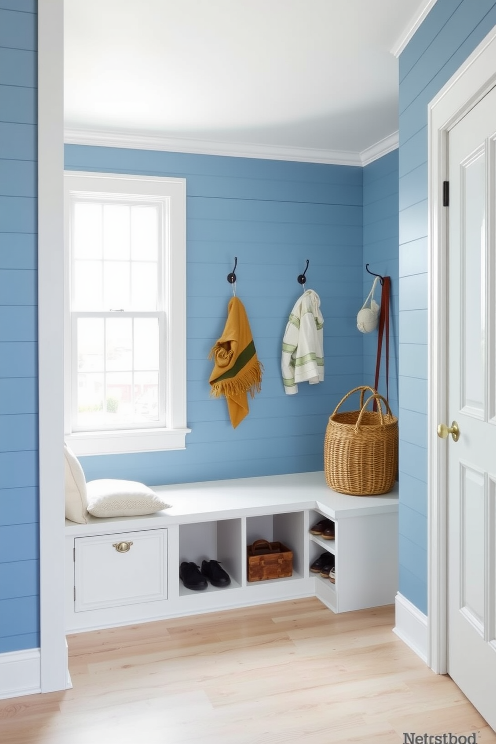 A coastal blue shiplap wall creates a serene backdrop in the mudroom, complemented by crisp white trim that enhances the fresh aesthetic. The space features a built-in bench with soft cushions and ample storage for shoes and outdoor gear, ensuring functionality meets style. Natural light floods the room through a large window, illuminating the light wood flooring and adding warmth to the cool color palette. Decorative hooks line the wall for easy access to jackets and bags, while a woven basket adds texture and organization to the space.