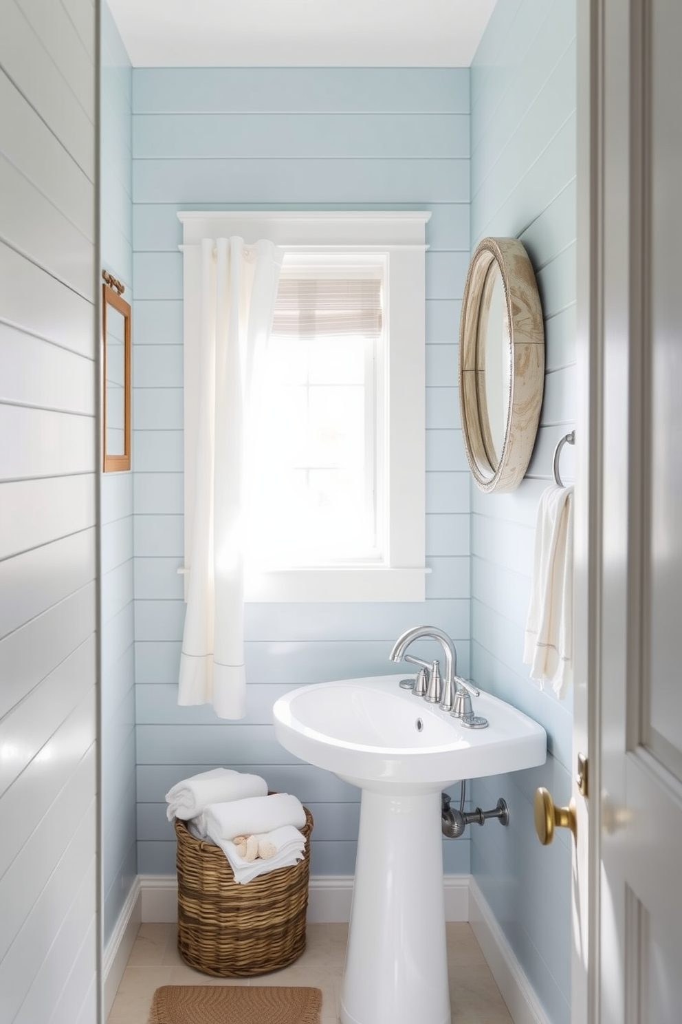 Light blue shiplap walls create a serene and inviting atmosphere in this coastal powder room. The space features a sleek white pedestal sink complemented by a round mirror with a driftwood frame, enhancing the beachy vibe. Natural light floods the room through a small window adorned with sheer white curtains. A woven basket sits in the corner, filled with plush towels and seashell accents, adding a touch of coastal decor.