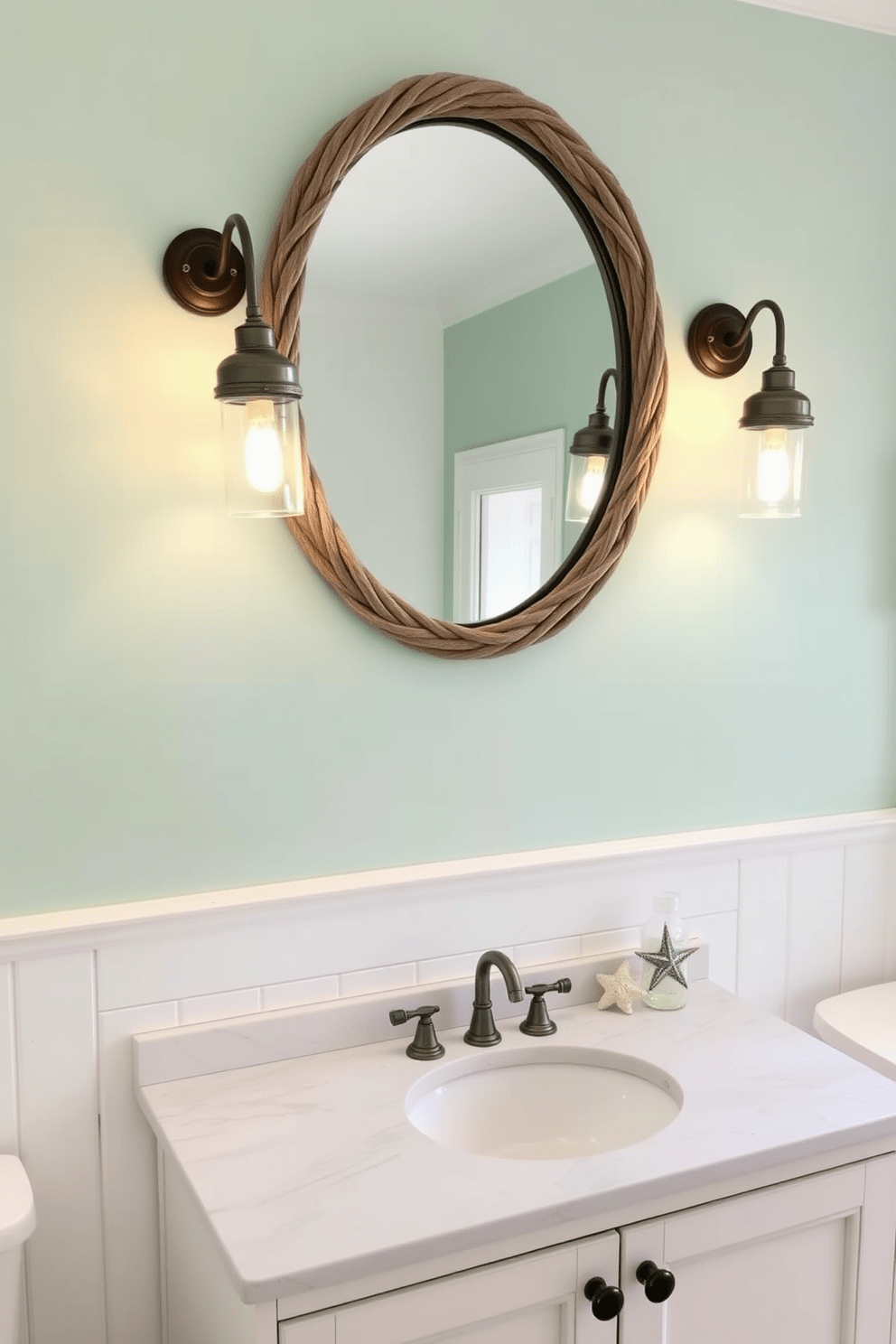 Sconces with a nautical theme illuminate the space, featuring rope accents and glass shades reminiscent of lanterns. The walls are painted in soft seafoam green, complemented by white shiplap paneling for a fresh coastal feel. The vanity is a distressed white wood with a marble top, adorned with seashell hardware. A large round mirror hangs above, framed in weathered driftwood, reflecting the natural light from the sconces.