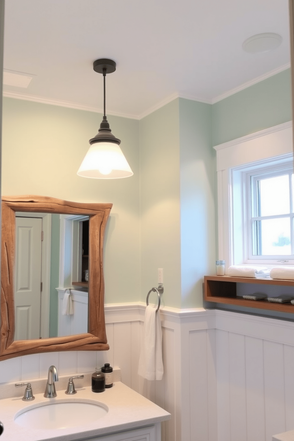 Layered lighting creates a cozy ambiance in a coastal bathroom design. Soft recessed lights illuminate the space while pendant fixtures add warmth above the vanity area. The walls are painted in a light seafoam green, complemented by white wainscoting for a fresh coastal feel. Natural wood accents, such as a driftwood mirror frame and shelving, enhance the beachy aesthetic.