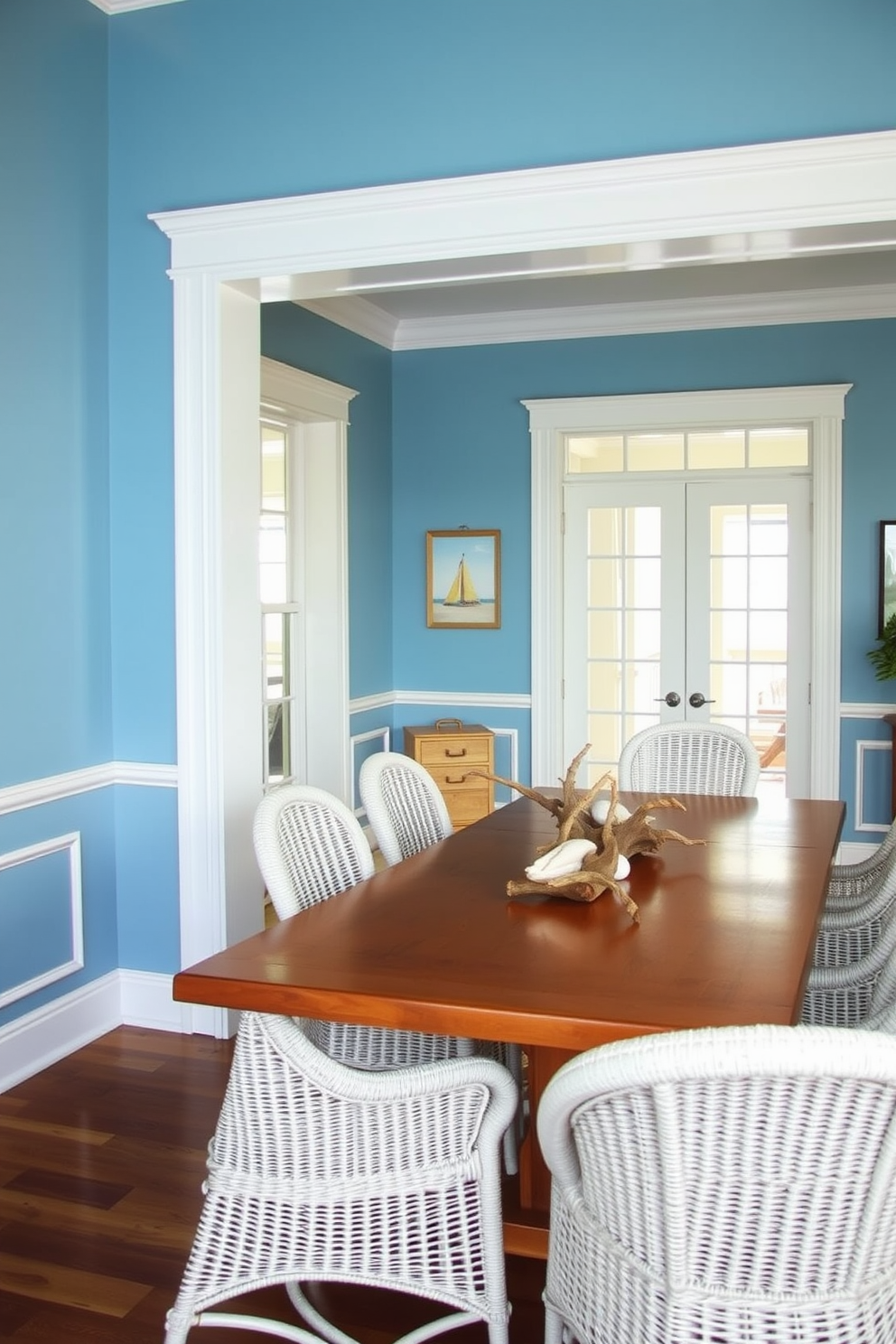 Light blue walls with white trim create a serene coastal atmosphere in the dining room. A large wooden table is surrounded by white wicker chairs, and a centerpiece of seashells and driftwood adds a natural touch.