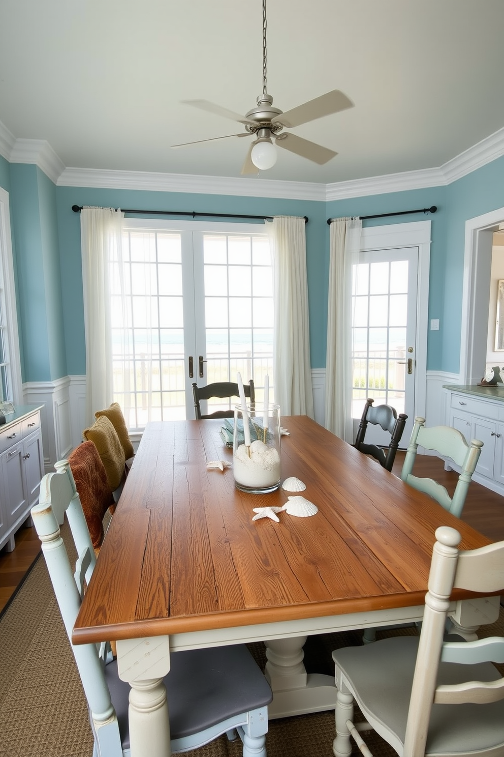 A coastal dining room featuring a large wooden table with a distressed finish surrounded by mismatched chairs. A glass vase filled with beach sand sits at the center of the table, complemented by seashells and candles for a relaxed ambiance. The walls are painted in soft blue tones, evoking the colors of the ocean. Large windows allow natural light to flood the space, with sheer white curtains gently billowing in the breeze.