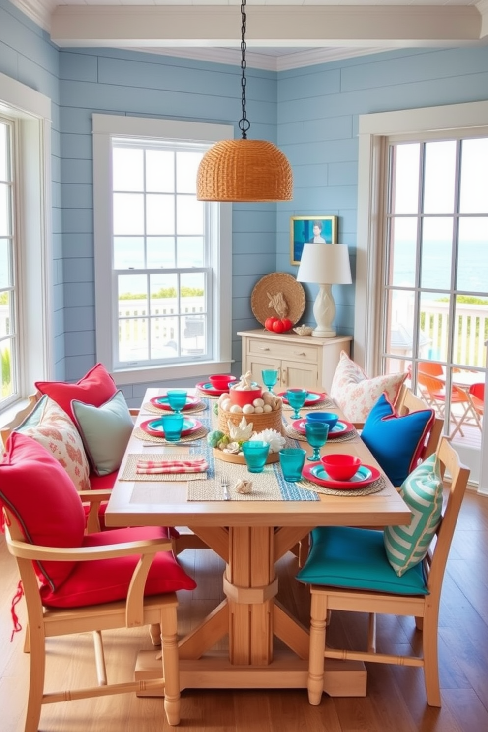 Brightly colored cushions are scattered across a light-colored wooden dining set, enhancing the inviting atmosphere of the coastal dining room. The walls are adorned with soft blue shiplap, and large windows offer a view of the ocean, allowing natural light to flood the space. The table is set with a mix of vibrant tableware, featuring coral and turquoise hues that reflect the coastal theme. Nautical decor elements, such as a woven centerpiece and seashell accents, complete the look, creating a relaxed yet stylish dining experience.