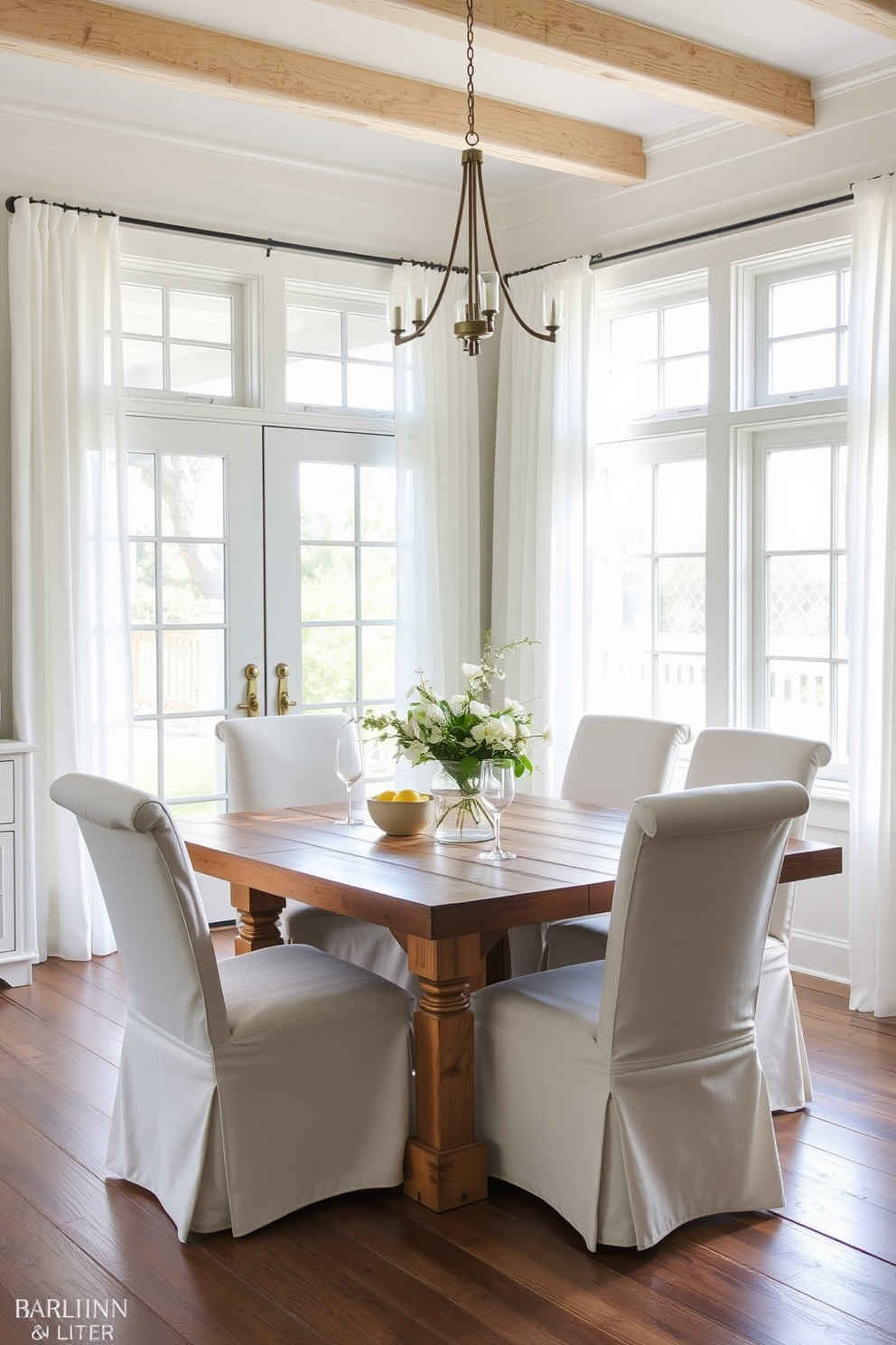 A bright coastal dining room featuring slipcovered chairs in light linen fabric. The table is made of reclaimed wood and is surrounded by soft, natural light coming through large windows adorned with sheer white curtains.