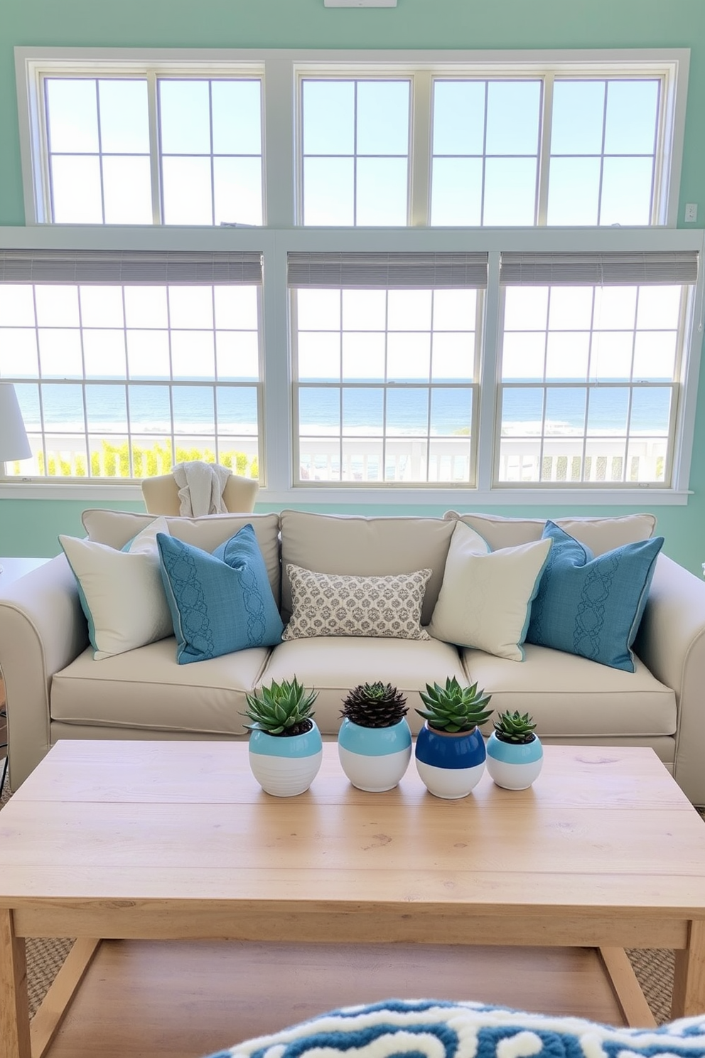 A coastal living room filled with natural light. The room features a soft beige sofa adorned with blue and white throw pillows, complemented by a light wood coffee table. Succulents in beach-themed pots are placed on the table, adding a touch of greenery. The walls are painted in a soft turquoise, and large windows offer a view of the ocean.