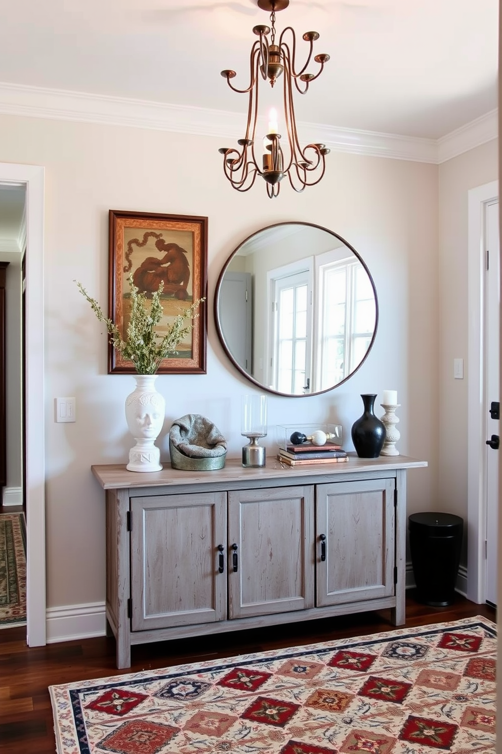 A contemporary foyer featuring vintage accents that create a warm and inviting atmosphere. The space includes a sleek console table with a distressed wood finish, adorned with a large round mirror and an eclectic assortment of decorative items. An elegant chandelier hangs from the ceiling, casting a soft glow over the area. The walls are painted in a soft neutral tone, complemented by a patterned area rug that adds texture and interest to the floor.