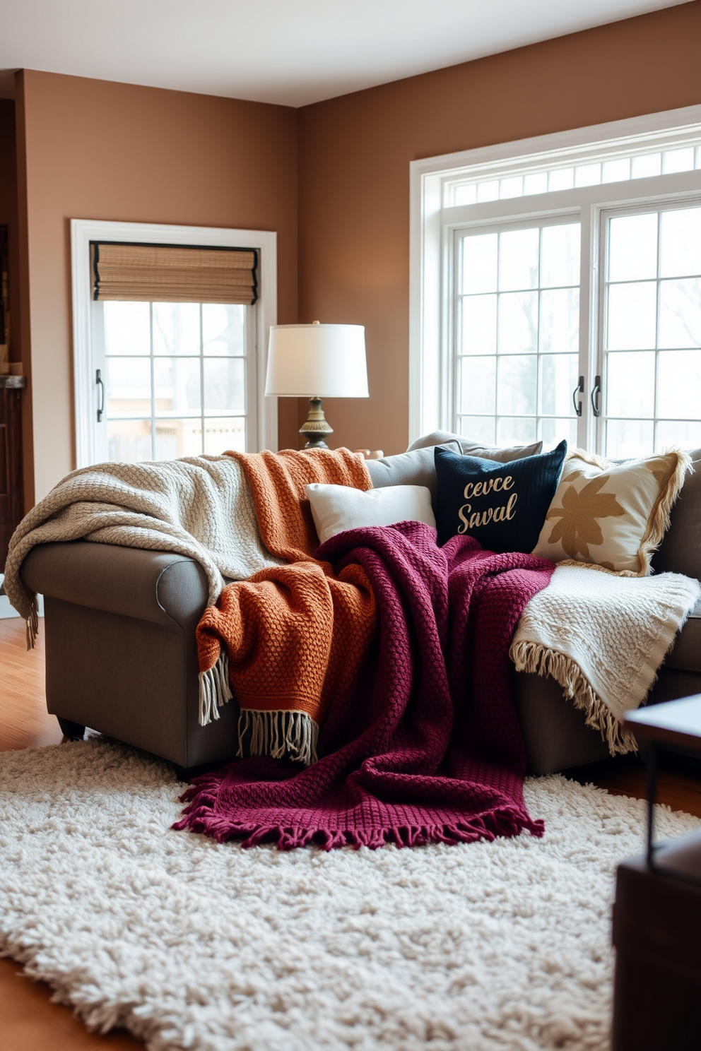 Cozy throw blankets are draped over a plush sectional sofa, inviting warmth and comfort. A soft area rug anchors the space, complementing the rich textures of the blankets and providing a perfect spot for relaxation. The walls are painted in a warm neutral tone, enhancing the inviting atmosphere of the living room. Natural light filters in through large windows, illuminating the space and highlighting decorative pillows that add pops of color to the cozy setting.