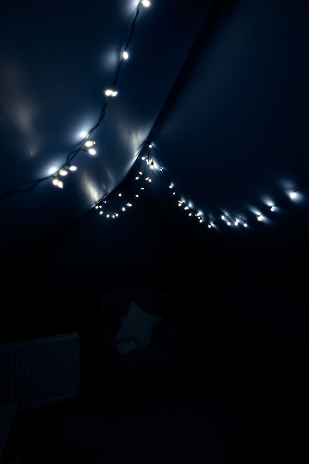 A dark attic room filled with mood lighting created by soft fairy lights strung across the ceiling. The walls are painted in a deep navy blue, and a cozy reading nook with a plush armchair is nestled under the slanted roof.