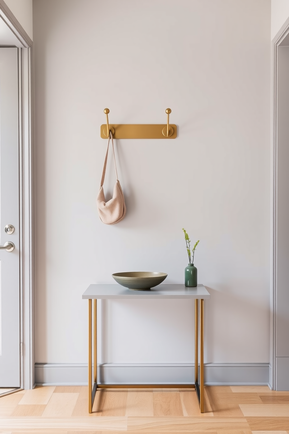 A stylish entrance foyer featuring elegant wall hooks for functional organization. The hooks are made of brushed brass and are mounted on a soft gray wall, providing a modern yet welcoming touch. A sleek console table sits beneath the hooks, adorned with a decorative bowl and a small potted plant. The floor is covered with a warm wooden finish, enhancing the inviting atmosphere of the space.