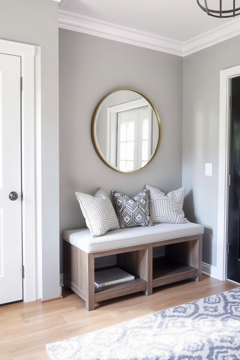 A chic bench with storage underneath is positioned against a wall in the entrance foyer. The bench features a soft upholstered seat in a neutral tone, complemented by decorative pillows for added comfort. Above the bench, a large round mirror reflects natural light, enhancing the space. The walls are painted in a soft gray hue, while a stylish area rug adds warmth and texture to the floor.