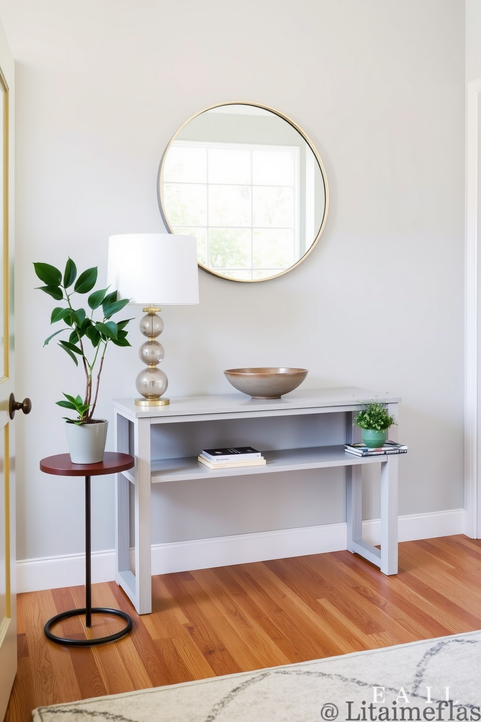 A welcoming entryway features a sleek console table against a soft gray wall, adorned with a stylish lamp and a decorative bowl. A small side table is placed nearby, showcasing a lush potted plant and a stack of art books for added decor. The floor is covered with a warm wood finish, complemented by a large area rug in neutral tones. A round mirror hangs above the console table, reflecting natural light from a nearby window, creating an inviting atmosphere.