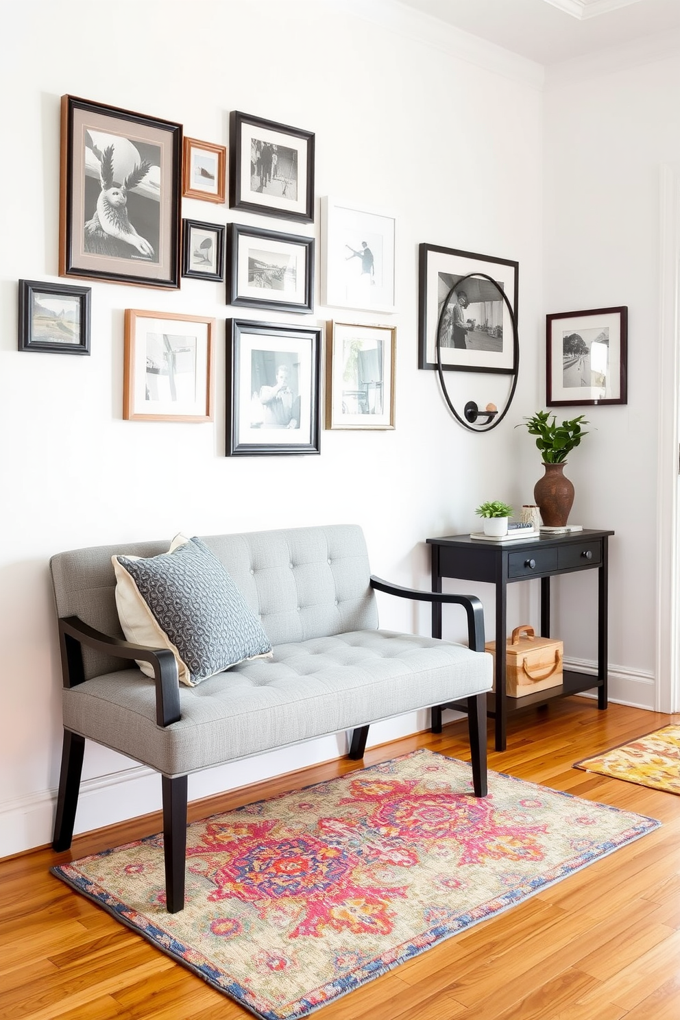 A welcoming entryway featuring a cozy bench upholstered in soft gray fabric. The bench is positioned against a light-colored wall adorned with a collection of framed artwork, creating an inviting atmosphere. A stylish console table sits beside the bench, topped with decorative items and a small potted plant. The floor is finished with warm hardwood, and a vibrant area rug adds a pop of color beneath the bench.