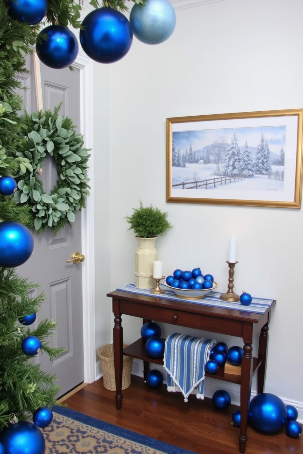 A welcoming entryway adorned with fresh greenery and blue ornaments creates a festive atmosphere. A beautifully arranged wreath made of pine and eucalyptus hangs on the door, while blue ornaments are tastefully scattered throughout the space. A small console table is decorated with a blue and white runner, topped with candles and a decorative bowl filled with ornaments. On the wall, a framed piece of art depicting winter scenes complements the holiday decor, enhancing the overall charm of the entryway.