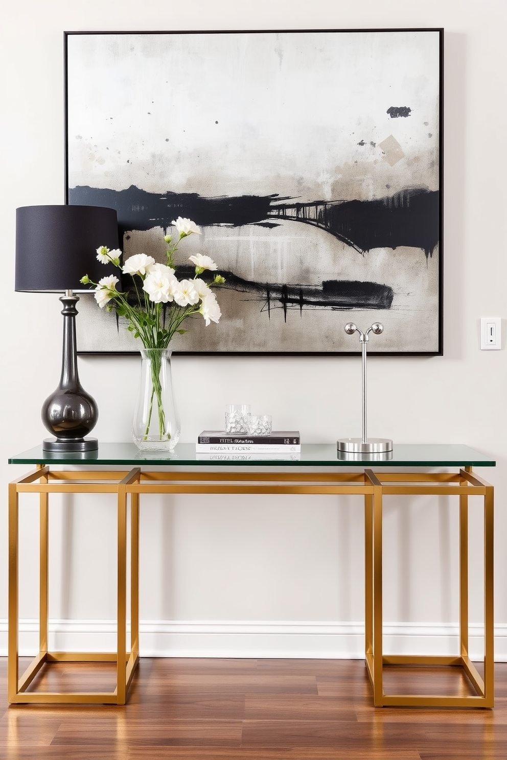 A sleek console table with metallic accents serves as a stunning focal point in the entryway. The table features a glass top and slender legs with a brushed gold finish, creating a modern and elegant look. Decorative elements such as a tall vase with fresh flowers and a stylish lamp are placed on the table. The wall behind the console is adorned with a large abstract painting that complements the metallic tones of the table.