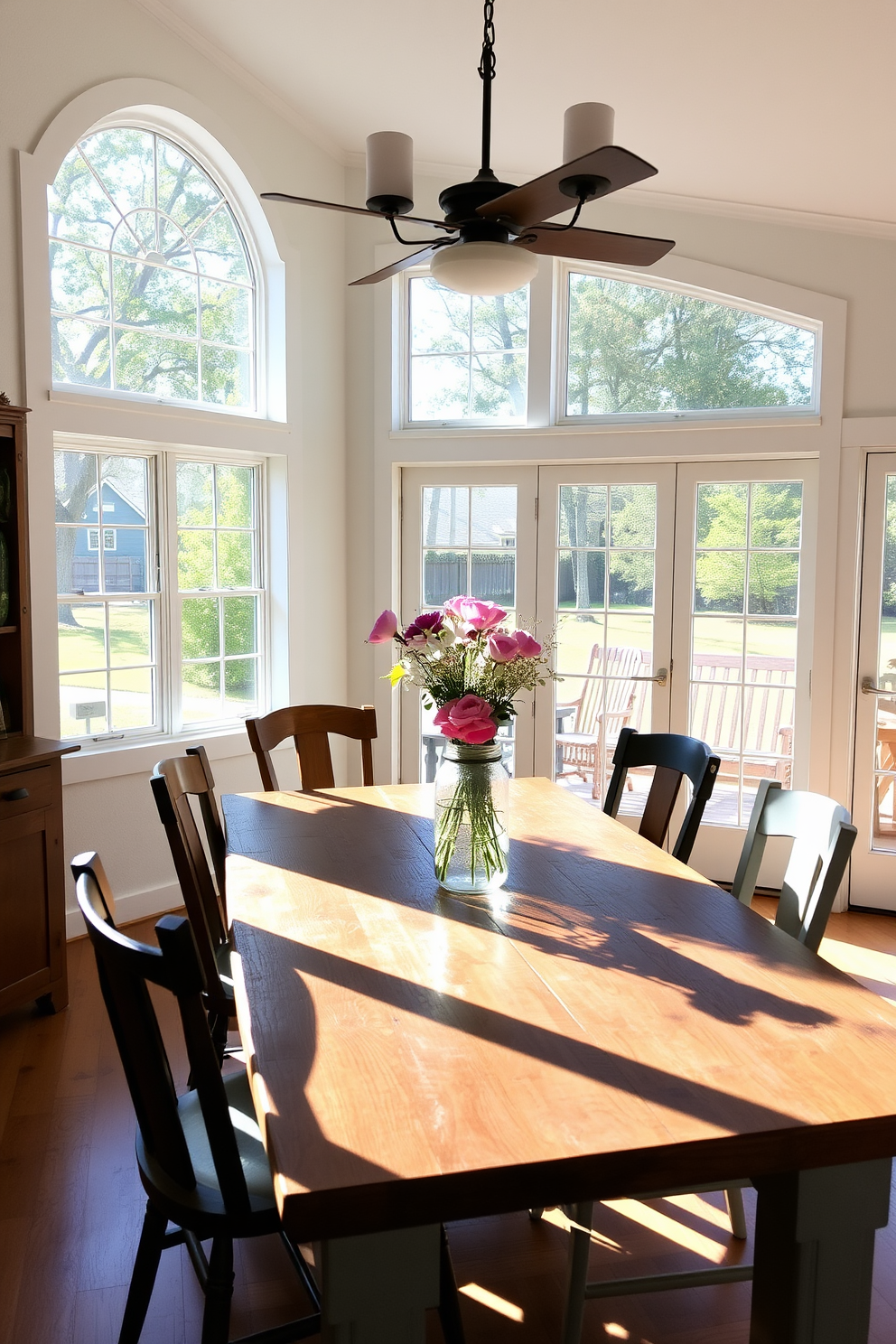 A bright farmhouse dining room filled with natural light streaming through large windows. The space features a rustic wooden table surrounded by mismatched chairs, with a centerpiece of fresh flowers in a mason jar.