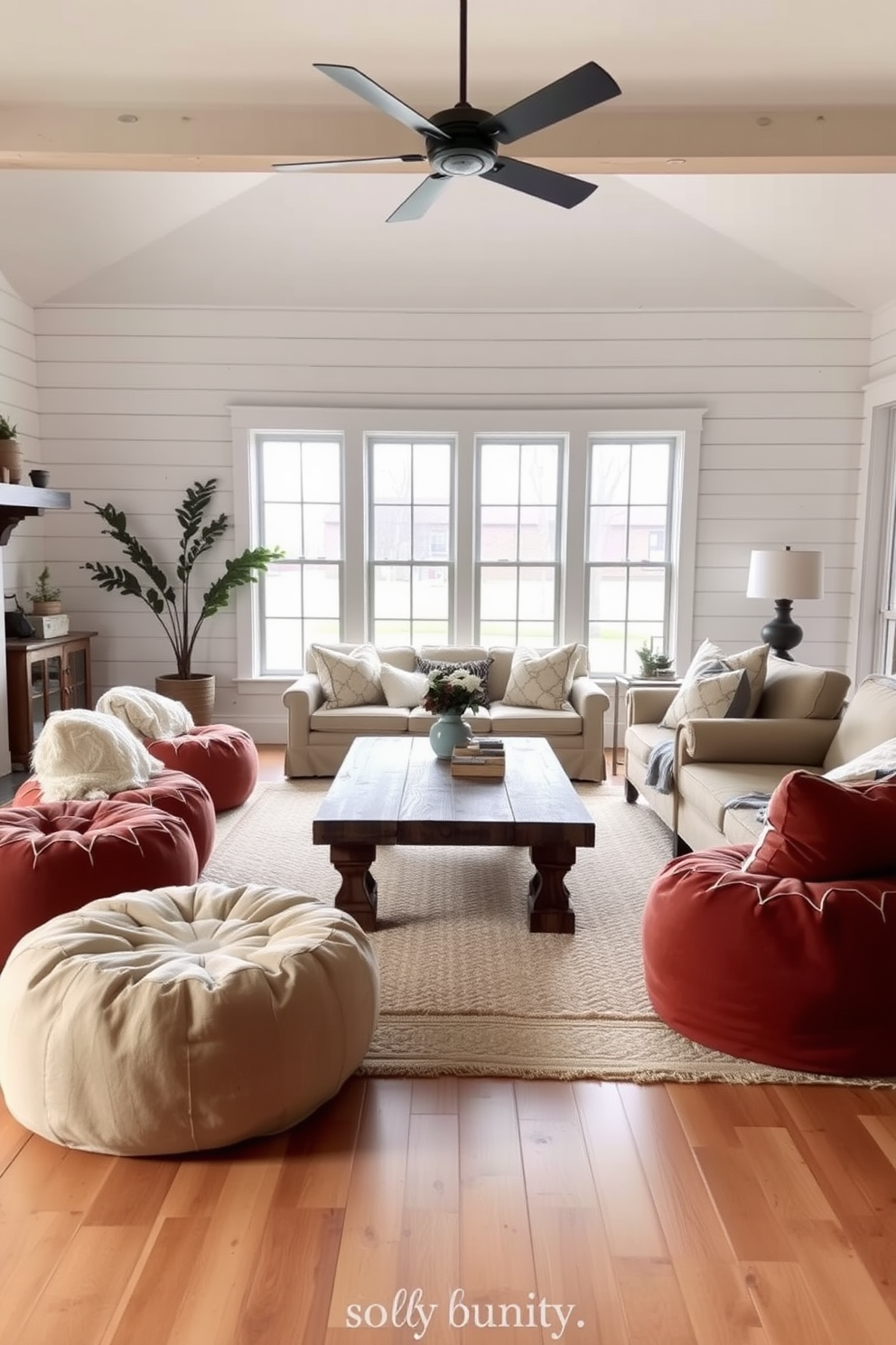 A cozy farmhouse living room featuring comfortable poufs arranged around a rustic wooden coffee table. The space is filled with natural light from large windows, and the walls are adorned with shiplap accents in soft white tones.