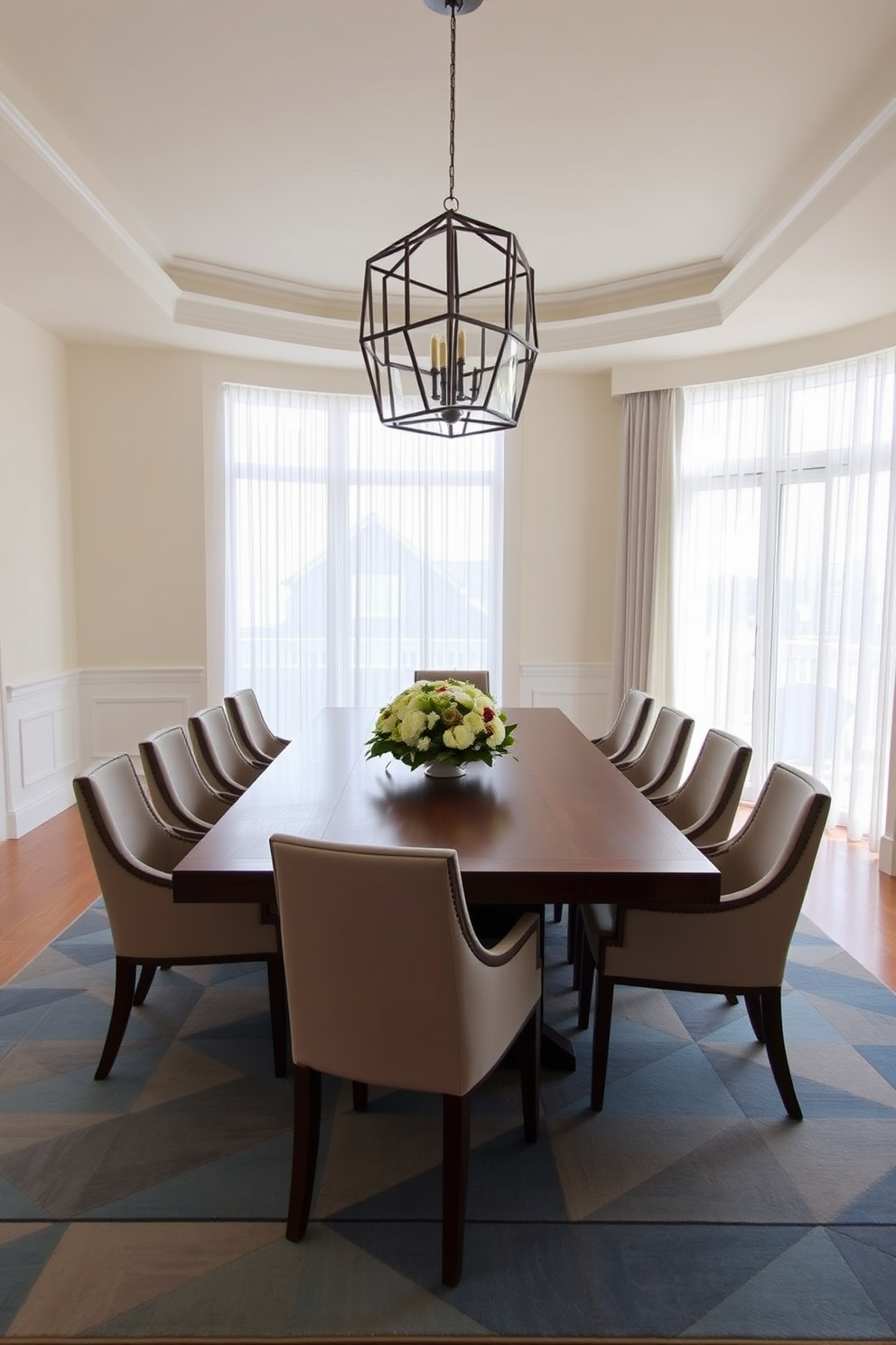 A formal dining room features a large rectangular table made of dark wood surrounded by upholstered chairs in a neutral fabric. The centerpiece of the table is an elegant floral arrangement, and a geometric rug in shades of blue and gray defines the dining area beneath it. The walls are painted a soft cream color, enhancing the light that pours in from large windows dressed with sheer curtains. Above the table, a modern chandelier with geometric shapes adds a touch of sophistication to the overall design.