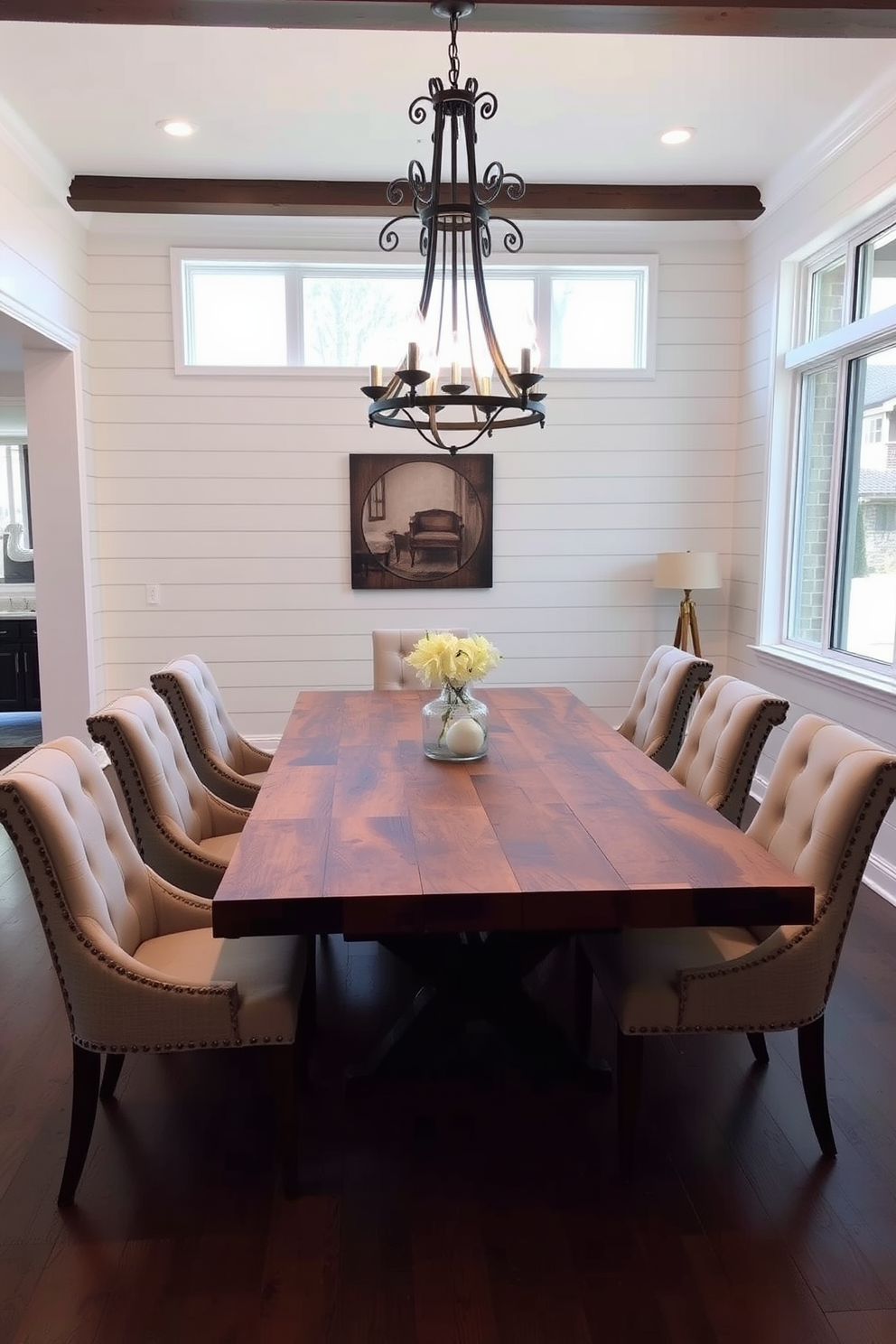 A rustic farmhouse table made of reclaimed wood sits at the center of a spacious dining room. Surrounding the table are elegant upholstered chairs in neutral tones, creating a warm and inviting atmosphere. Above the table, a vintage chandelier with wrought iron accents casts a soft glow. The walls are adorned with shiplap paneling, and a large window allows natural light to flood the room, enhancing the chic decor.