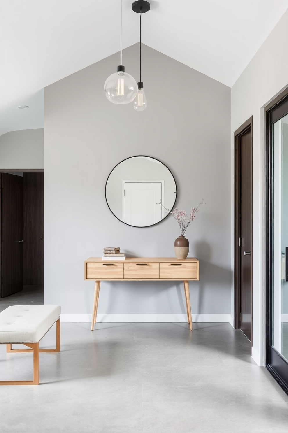 A modern foyer with sleek lines and a minimalist aesthetic. The space features a light wood console table against a backdrop of soft gray walls, complemented by a large round mirror above it. To the left, a stylish bench upholstered in neutral fabric provides a comfortable seating area. A statement pendant light hangs from the ceiling, casting a warm glow over the polished concrete floor.