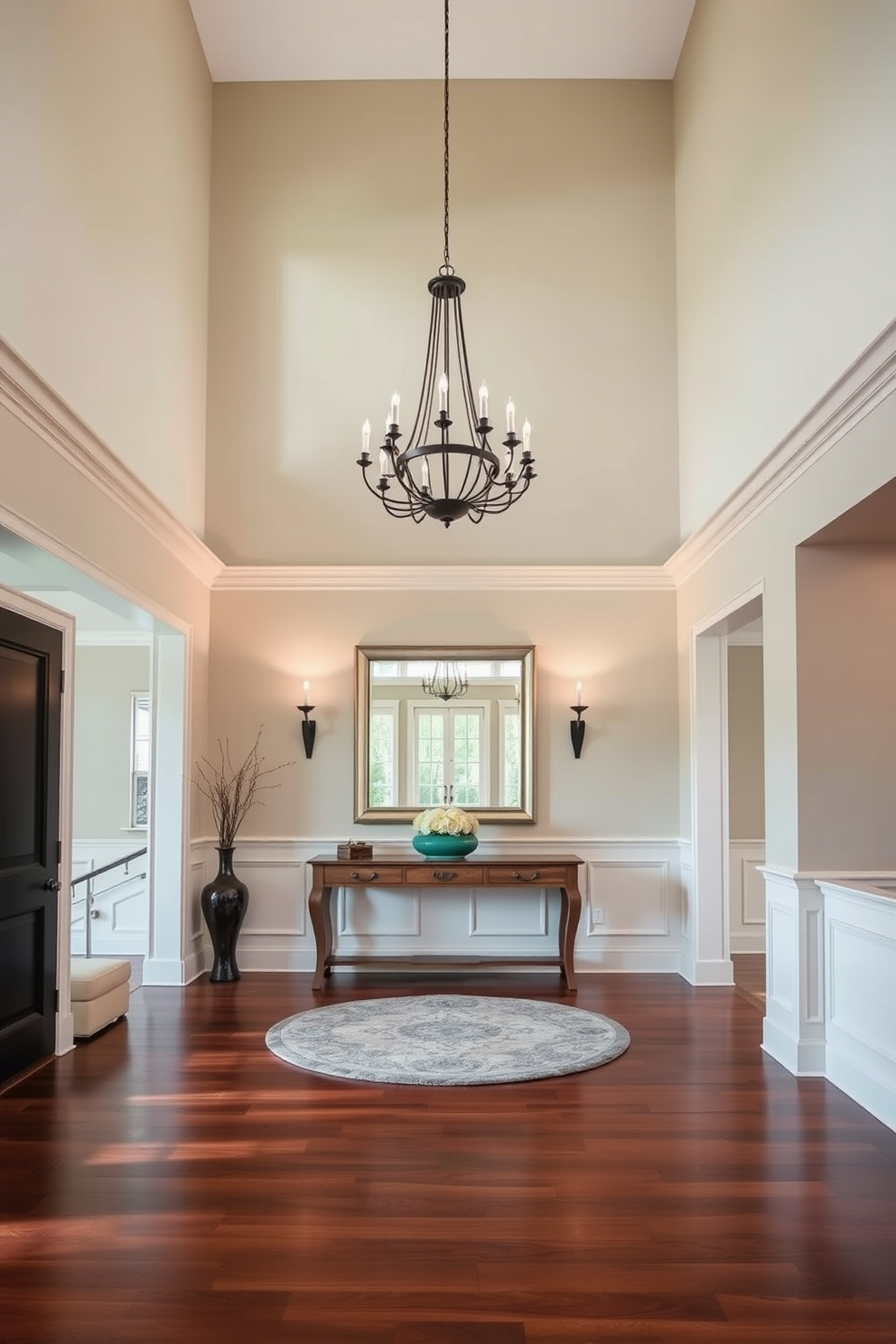 A grand foyer with high ceilings and an elegant chandelier hanging from the center. The walls are adorned with wainscoting painted in a soft cream color, and a large console table is placed against one wall. The floor features polished hardwood in a rich mahogany tone, complemented by a plush area rug in muted tones. A pair of stylish sconces flanks a large mirror above the console, creating a warm and inviting atmosphere.