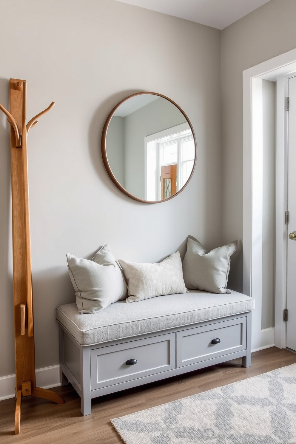 A chic bench with storage underneath is positioned against the wall in the foyer. The bench is upholstered in a soft gray fabric, complemented by decorative pillows in muted tones. Above the bench, a large round mirror reflects natural light, enhancing the space's openness. To one side, a stylish coat rack made of wood adds functionality while maintaining a cohesive design aesthetic.