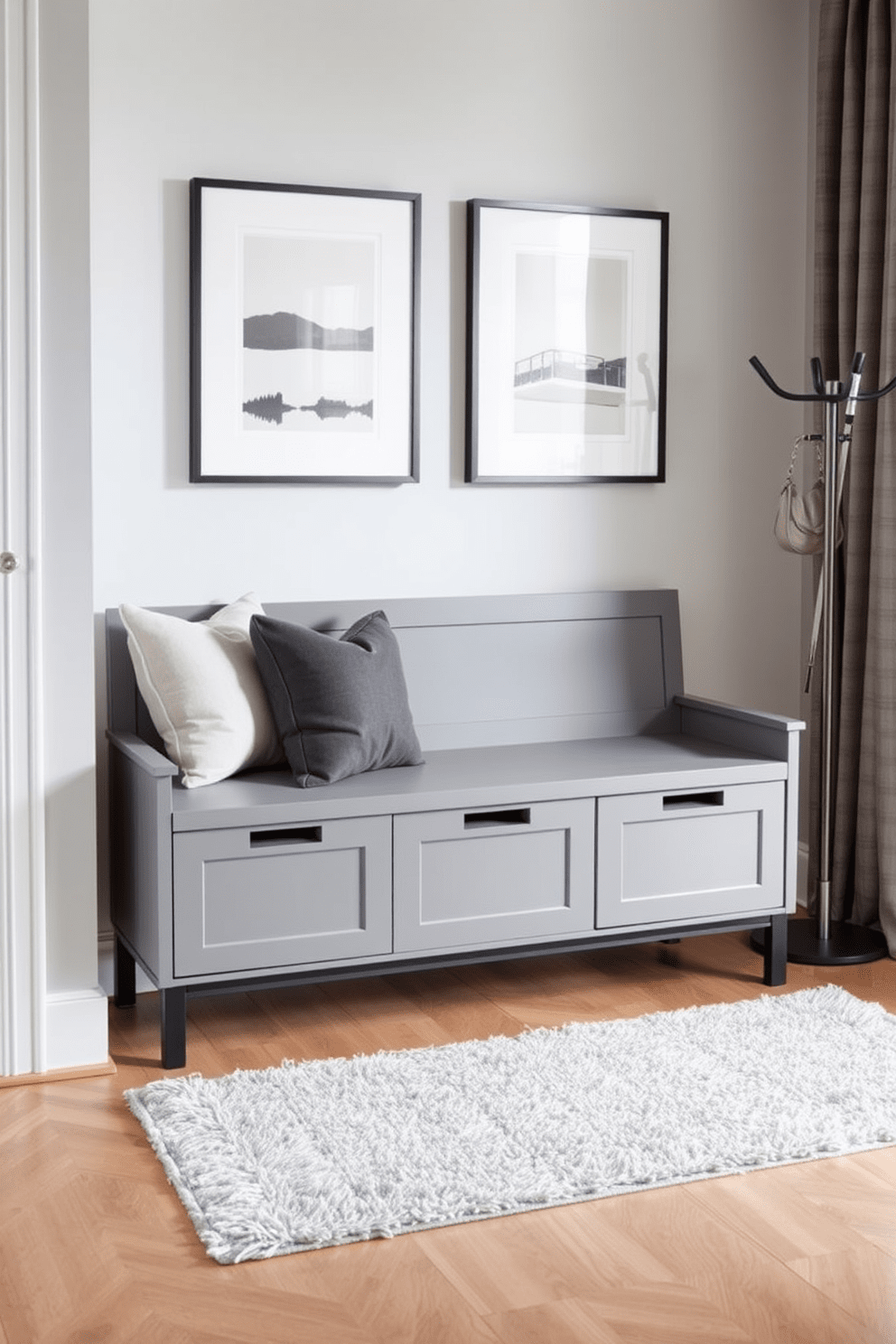 A stylish entryway featuring a gray bench with built-in storage compartments. The walls are adorned with minimalist artwork, and a soft rug lies beneath the bench, adding warmth to the space. The bench is complemented by decorative pillows in varying shades of gray. A sleek coat rack stands nearby, providing a functional yet elegant touch to the apartment design.