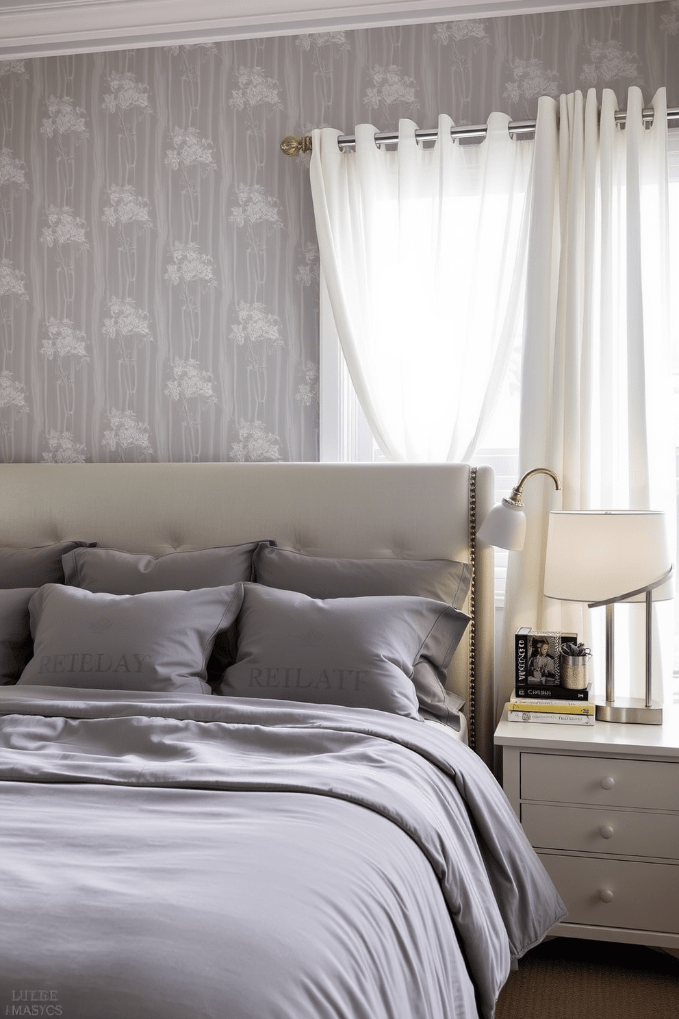 A serene gray bedroom featuring wallpaper with subtle floral patterns. The room is adorned with a plush gray bedspread and matching pillows, creating a calming atmosphere. Natural light filters through sheer white curtains, illuminating the space. A stylish nightstand holds a modern lamp and a few decorative books, enhancing the elegant design.