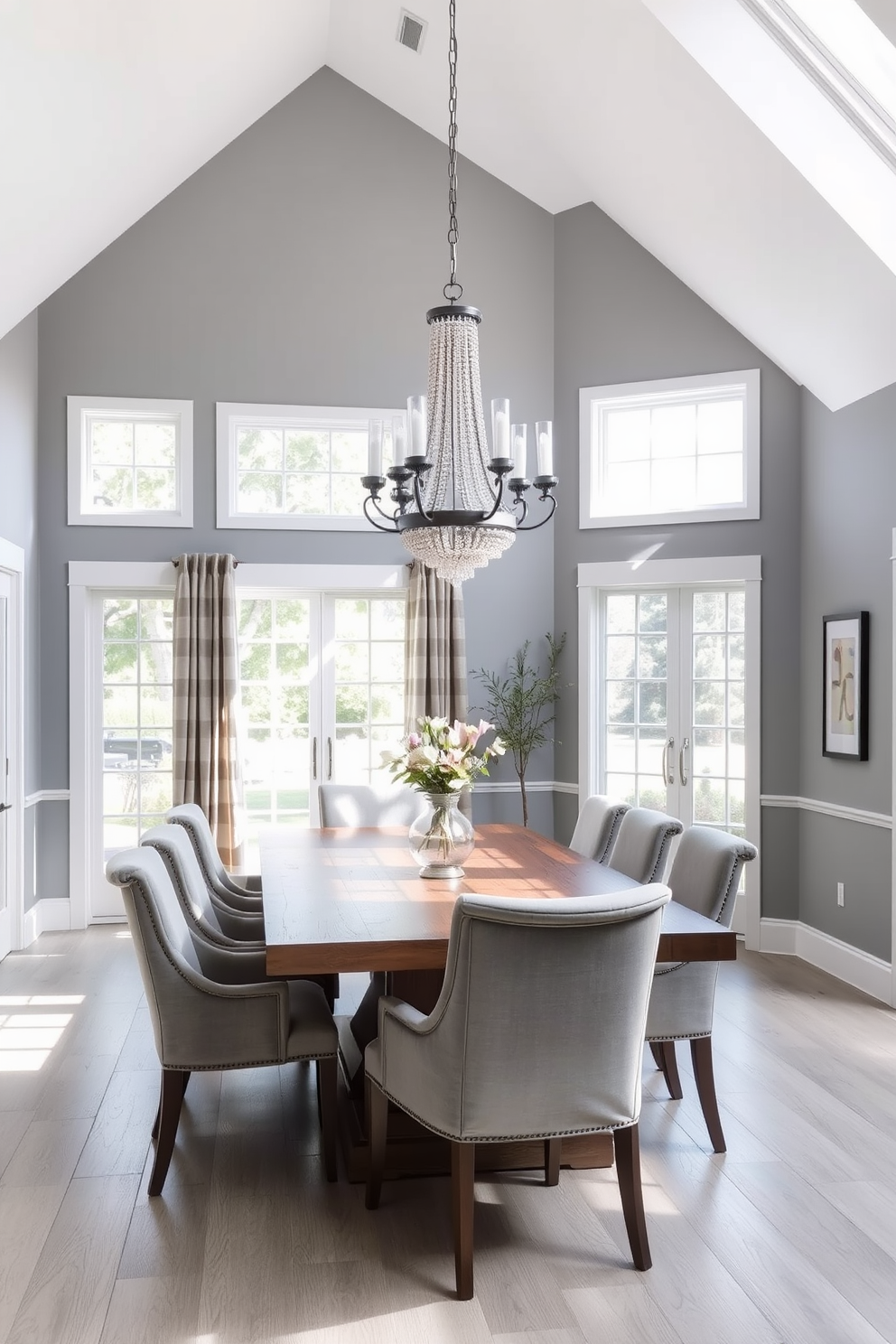 A stylish gray dining room filled with natural light. The space features a large wooden dining table surrounded by upholstered chairs in a soft gray fabric. Floor-to-ceiling windows allow sunlight to stream in, illuminating the room. A statement chandelier hangs above the table, adding a touch of elegance to the ambiance.