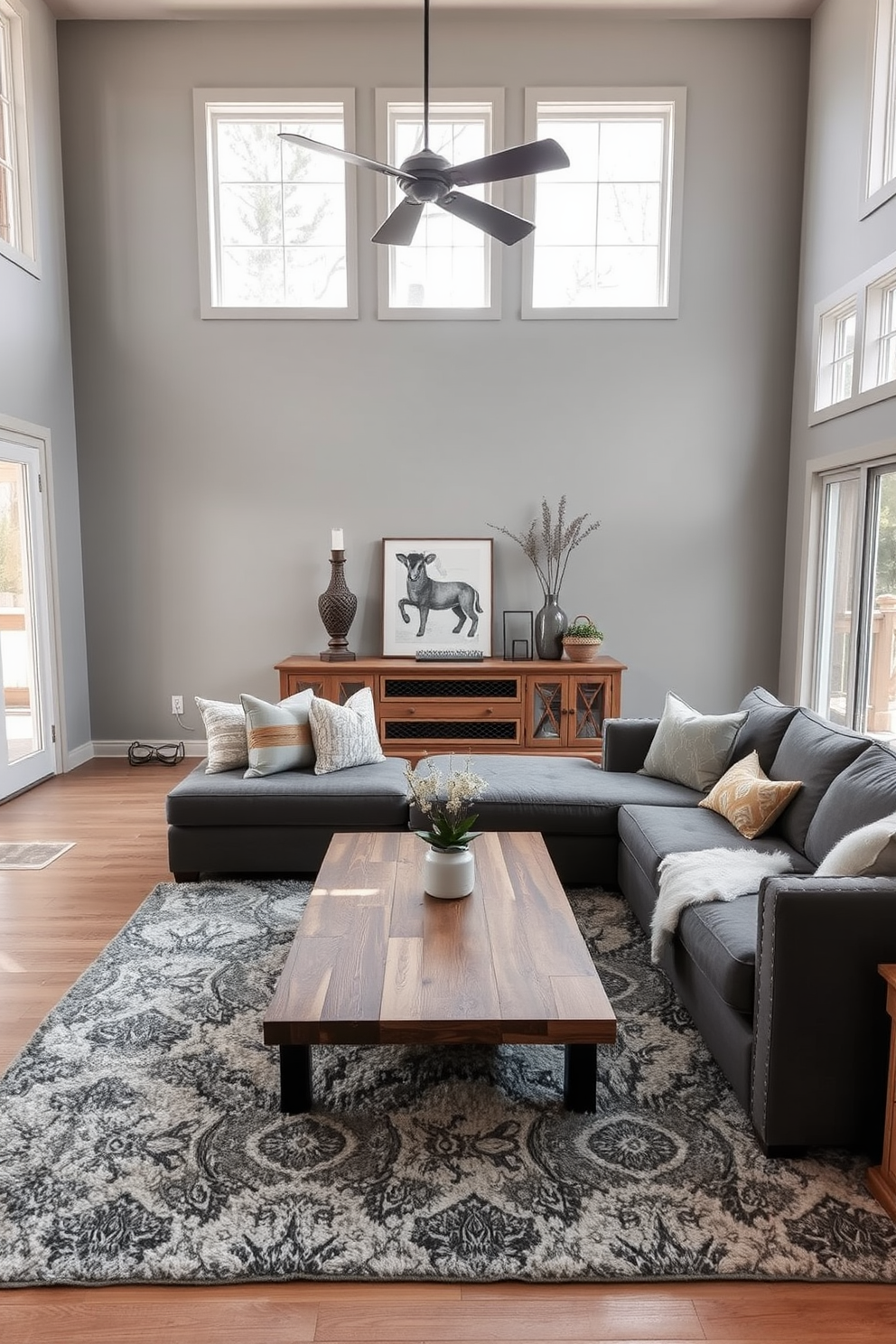 A cozy family room featuring layered gray tones complemented by natural wood accents. The walls are painted in a soft gray, and a large sectional sofa in a darker gray fabric invites relaxation. A reclaimed wood coffee table sits at the center, surrounded by plush area rugs that add texture. Floor-to-ceiling windows allow natural light to flood the space, enhancing the warm ambiance. Decorative throw pillows in varying shades of gray and a few vibrant accents create visual interest. A stylish media console made of natural wood adds functionality while maintaining the room's aesthetic.