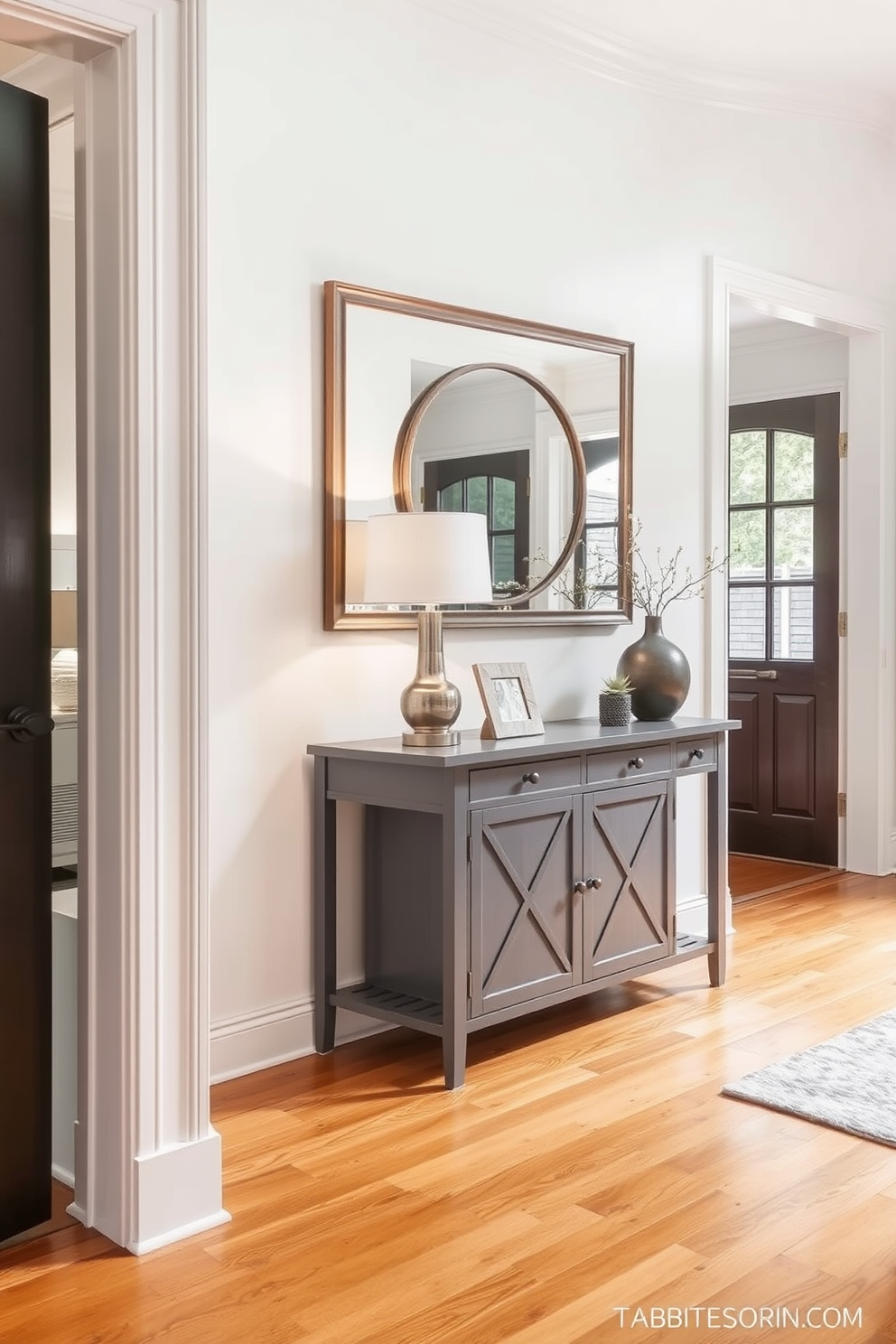 A stylish gray entryway table stands against the wall, adorned with decorative items such as a sleek lamp and a small potted plant. The walls are painted in a soft white hue, creating a bright and inviting atmosphere in the foyer. The floor features elegant hardwood that complements the gray tones of the table. A large mirror hangs above the table, reflecting light and adding depth to the space.