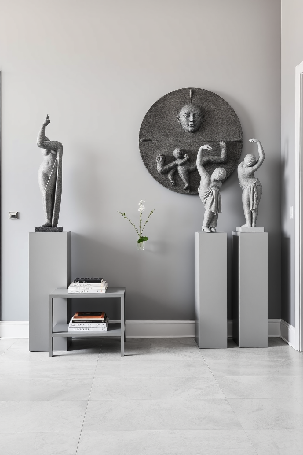 Artistic gray sculptures serve as striking focal points in a modern foyer. The walls are painted in a soft gray tone, complementing the elegant sculptures displayed on minimalist pedestals. A sleek console table in a matching gray finish sits against one wall, adorned with a few curated art books and a small potted plant. The flooring features large light gray tiles, creating a seamless and sophisticated look throughout the space.