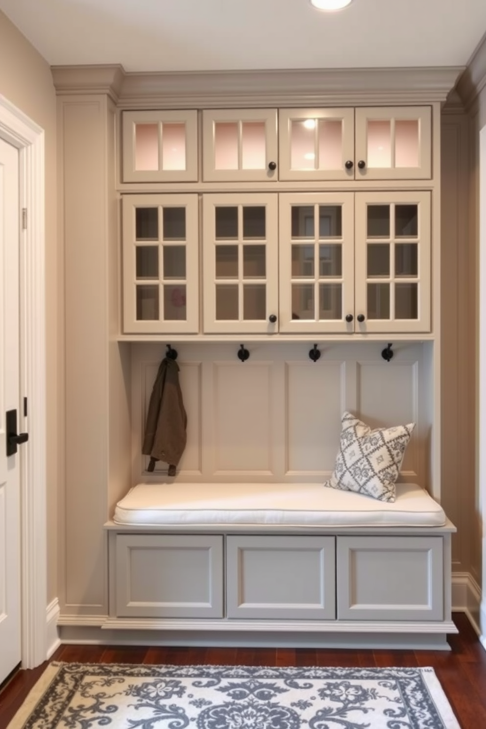 Light gray cabinets with glass fronts create an elegant and functional mudroom space. The room features a built-in bench with plush cushions and hooks for hanging coats, complemented by a stylish area rug.